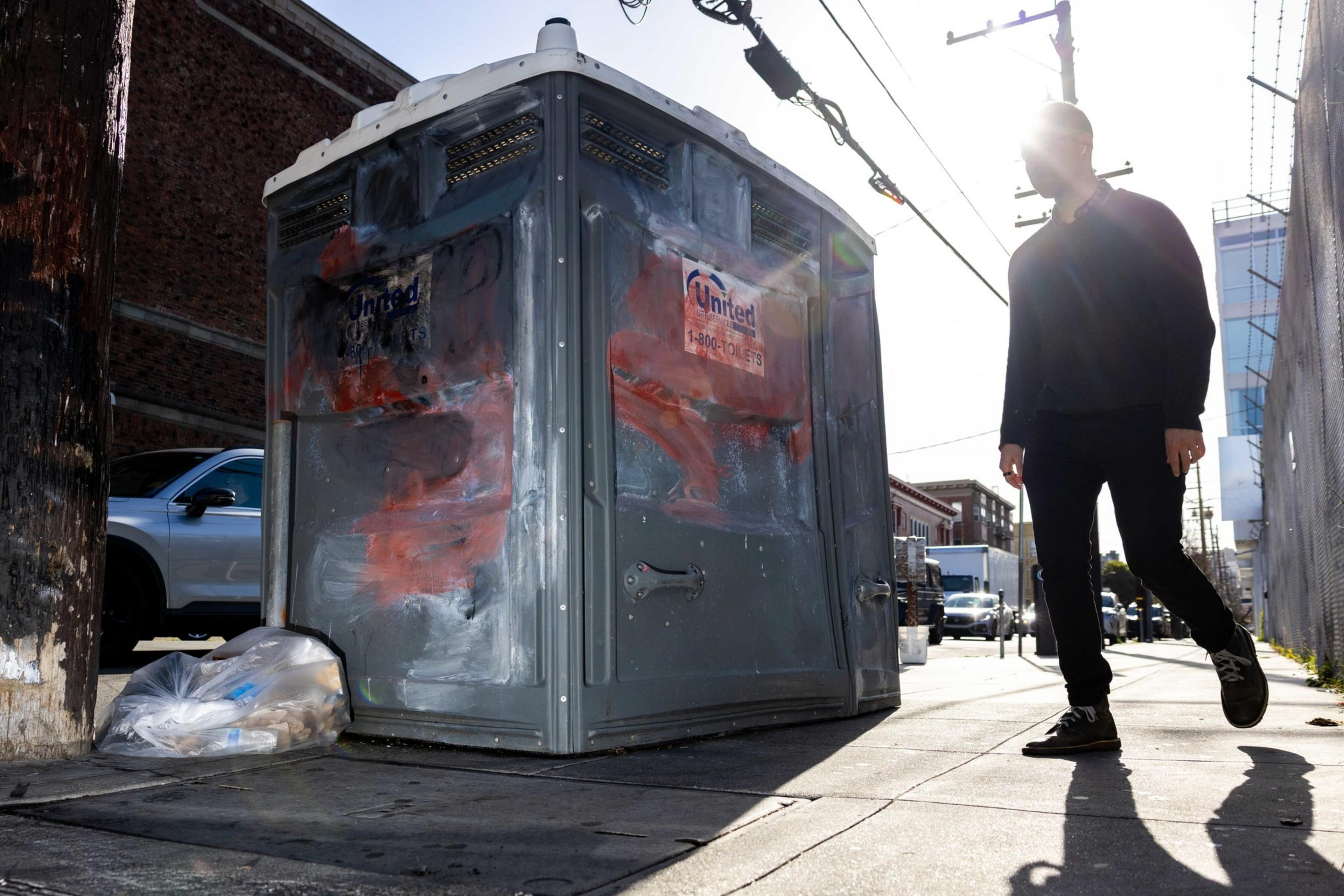 A person walks past a graffiti-marked portable toilet on a city sidewalk. The sun shines brightly, casting the person's shadow on the ground. A trash bag is nearby.