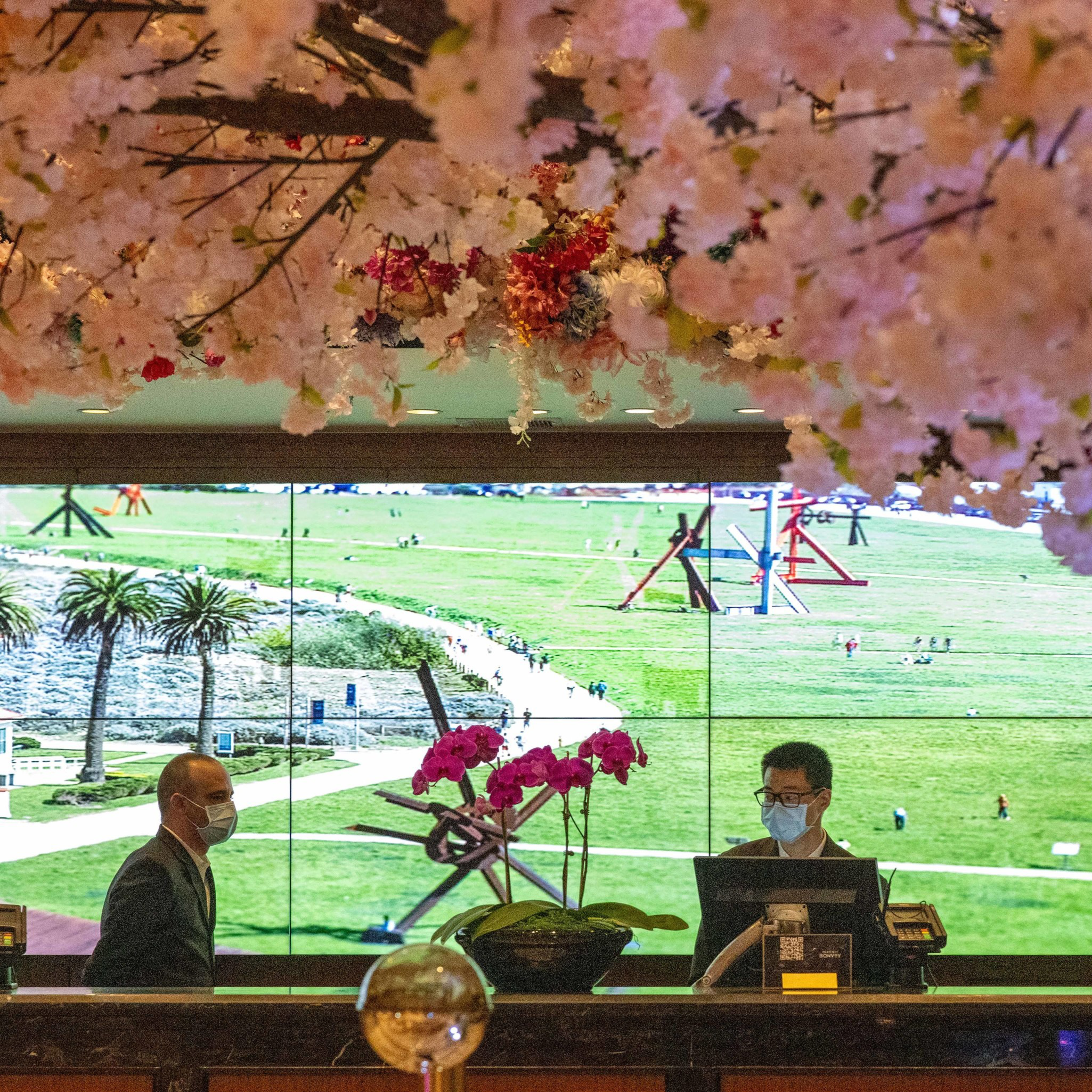 Two masked men stand behind a counter; above them are hanging pink flowers. An expansive green park background is visible, adorned with sculptures.