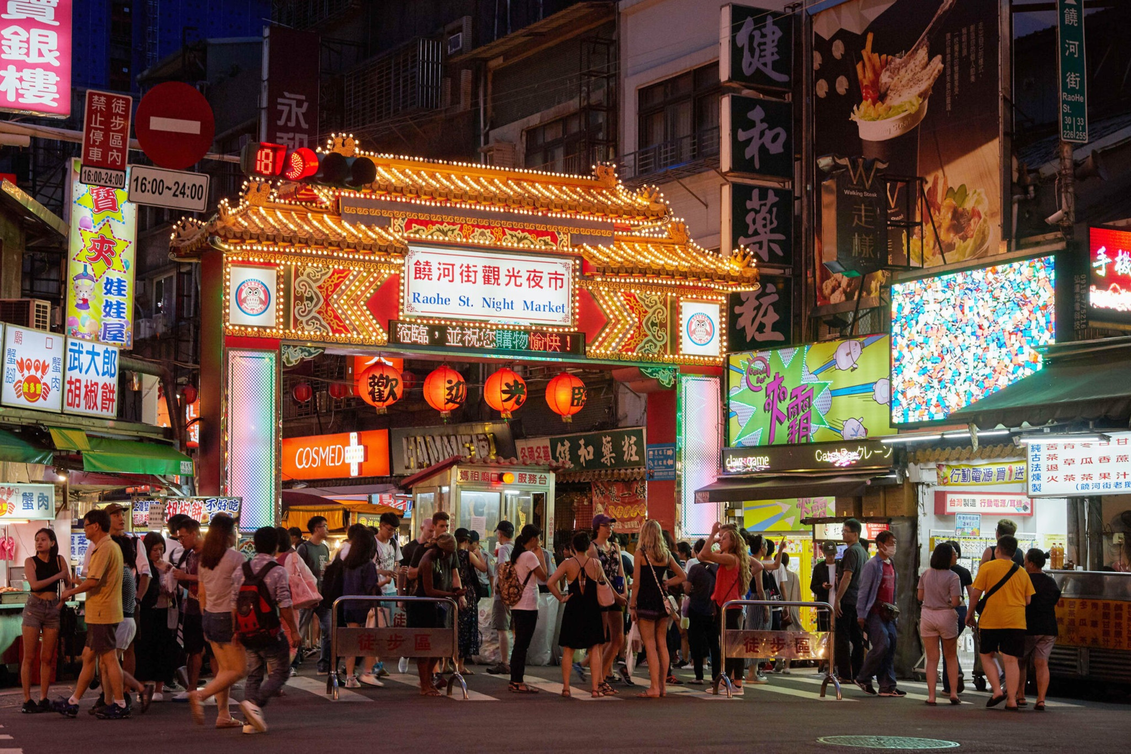 A brightly lit entrance to Raohe St. Night Market is crowded with people walking and standing, surrounded by colorful signs and street vendors.