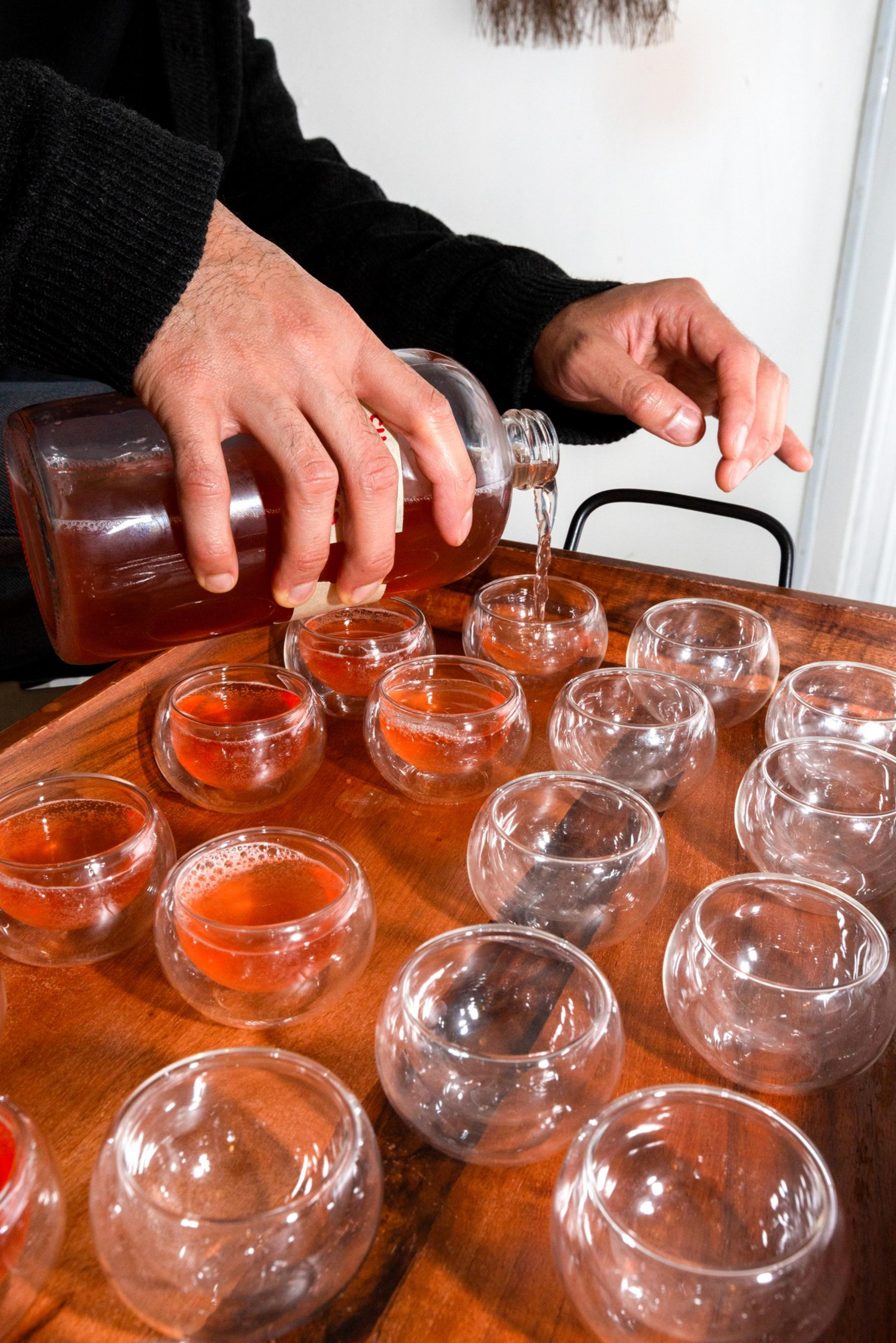 A person wearing a black sweater pours tea from a glass bottle into small, round glass cups arranged on a wooden tray.