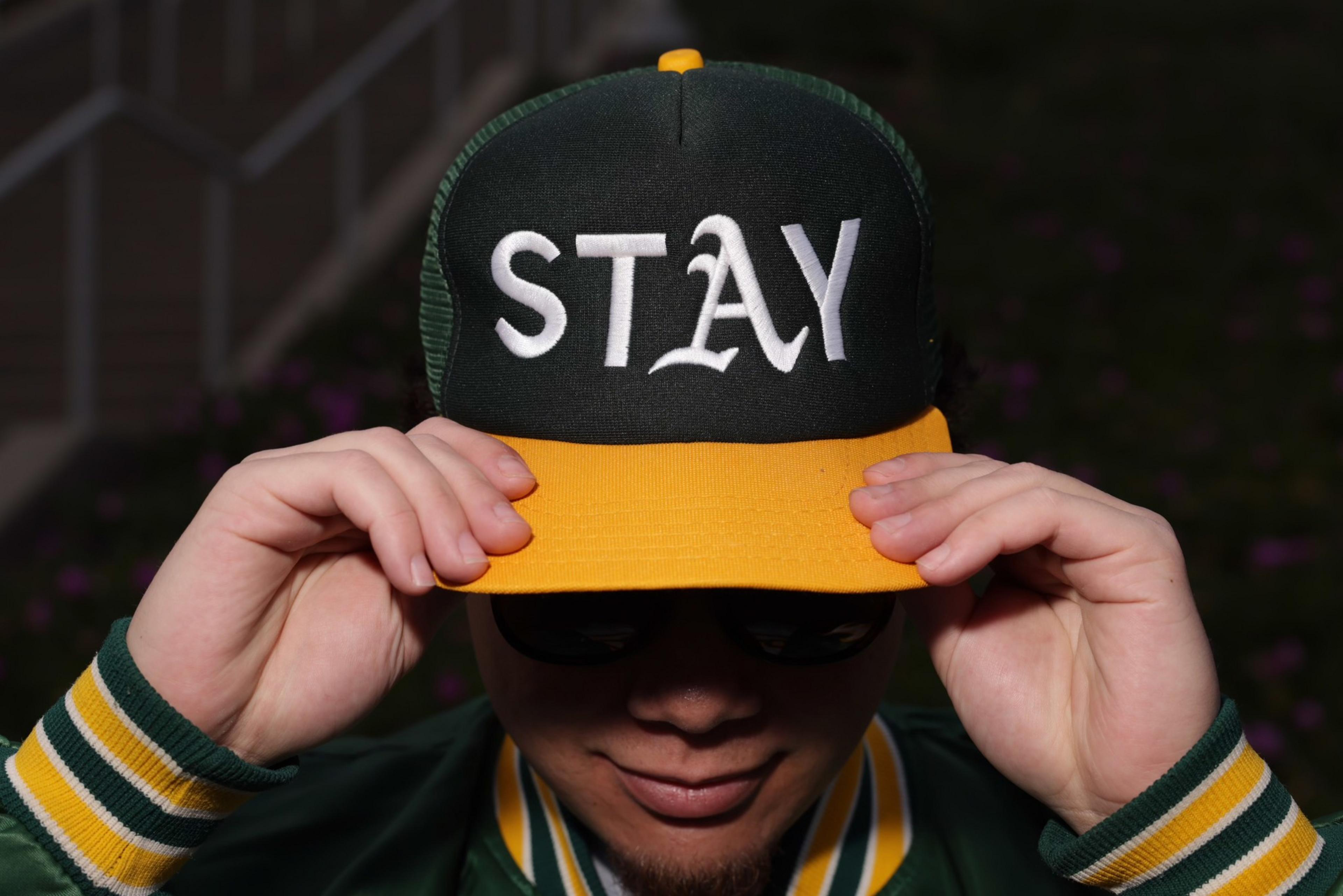 A person wearing a green and yellow cap with &quot;STAY&quot; written on it adjusts the brim. They are also wearing sunglasses and a green striped jacket.