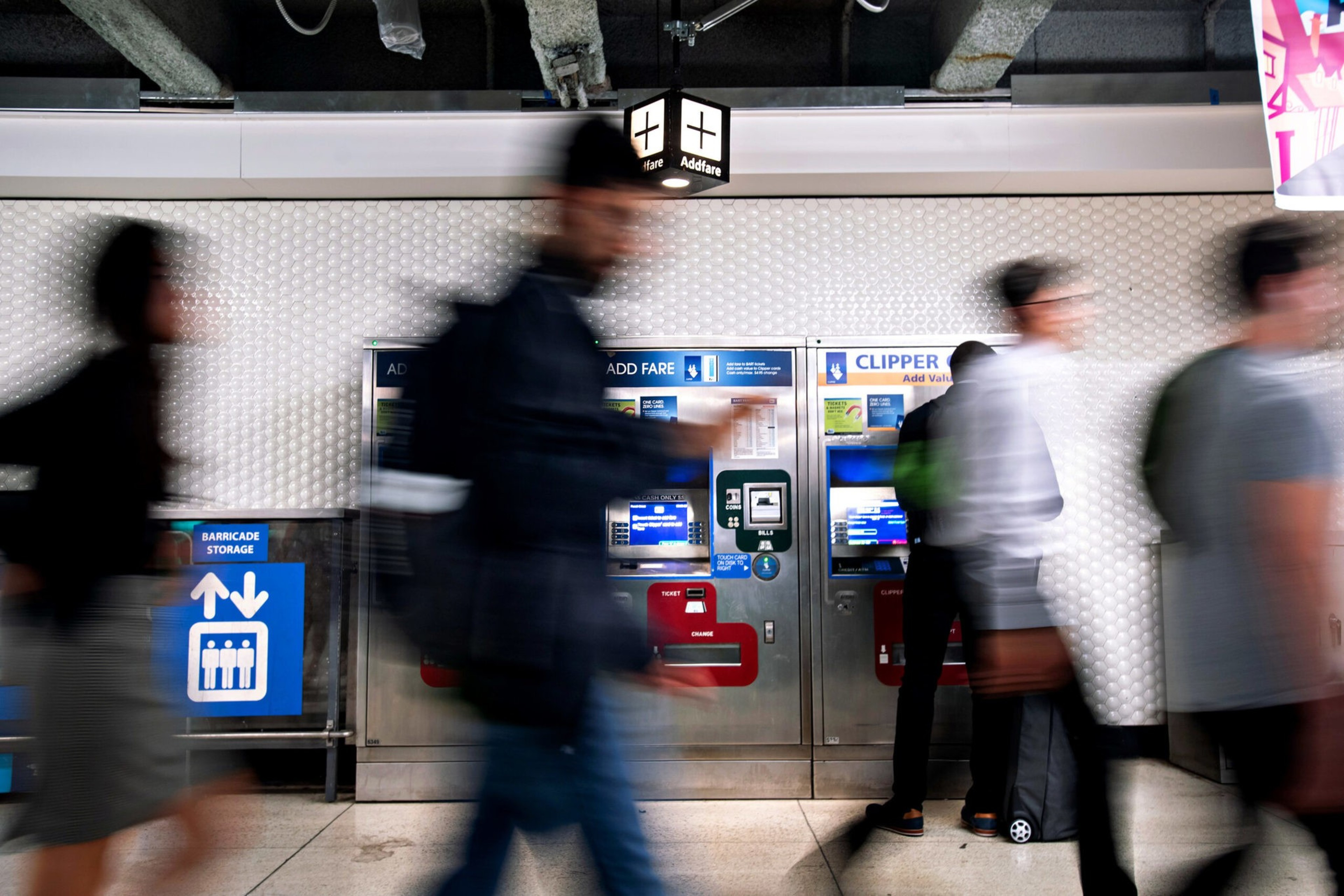 People walk past fare vending machines inside a transit station, with some individuals blurry from motion and a sign indicating "Add Fare" overhead.