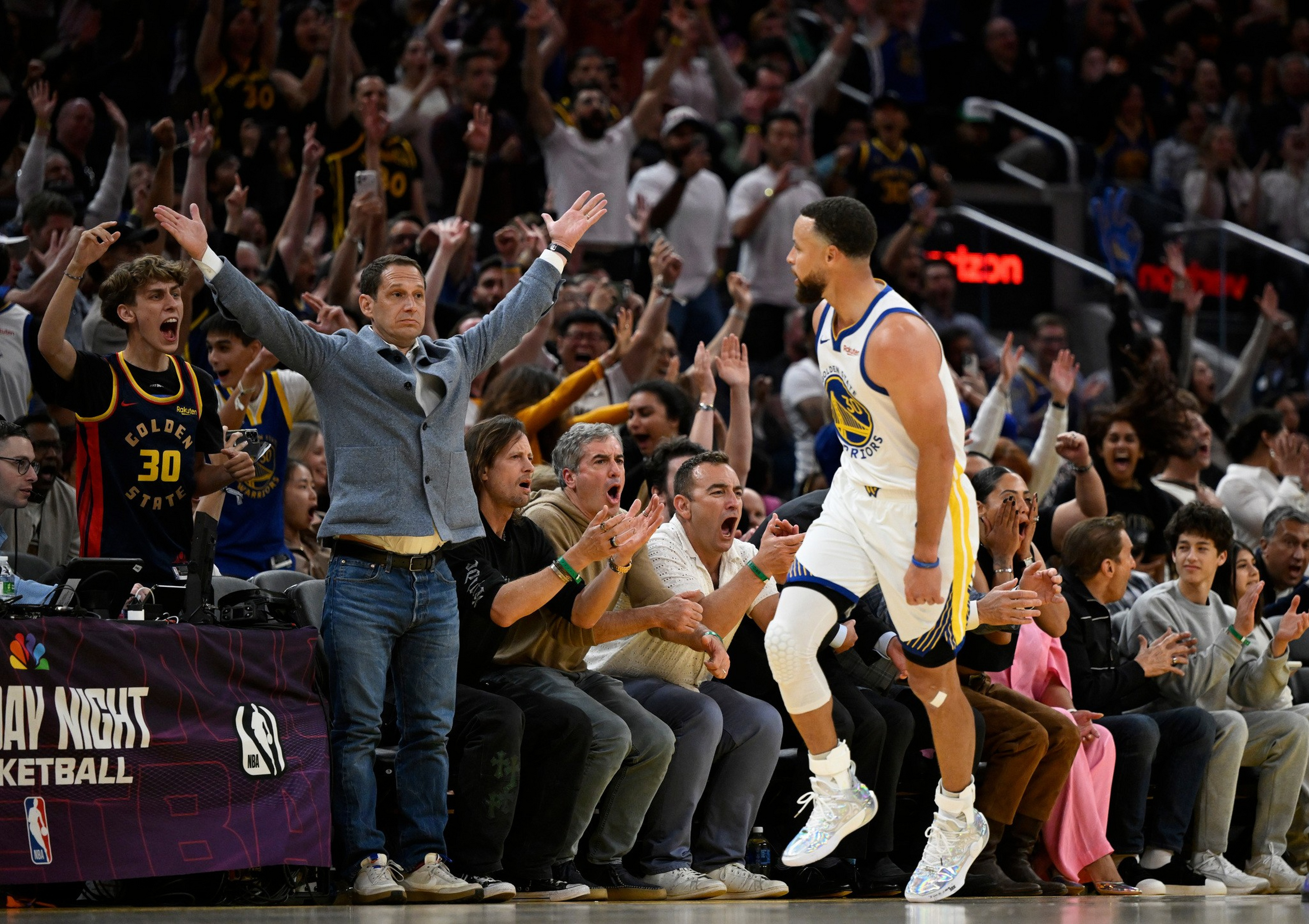 A Golden State Warriors player walks confidently past an excited crowd and coach celebrating a key moment during a basketball game.