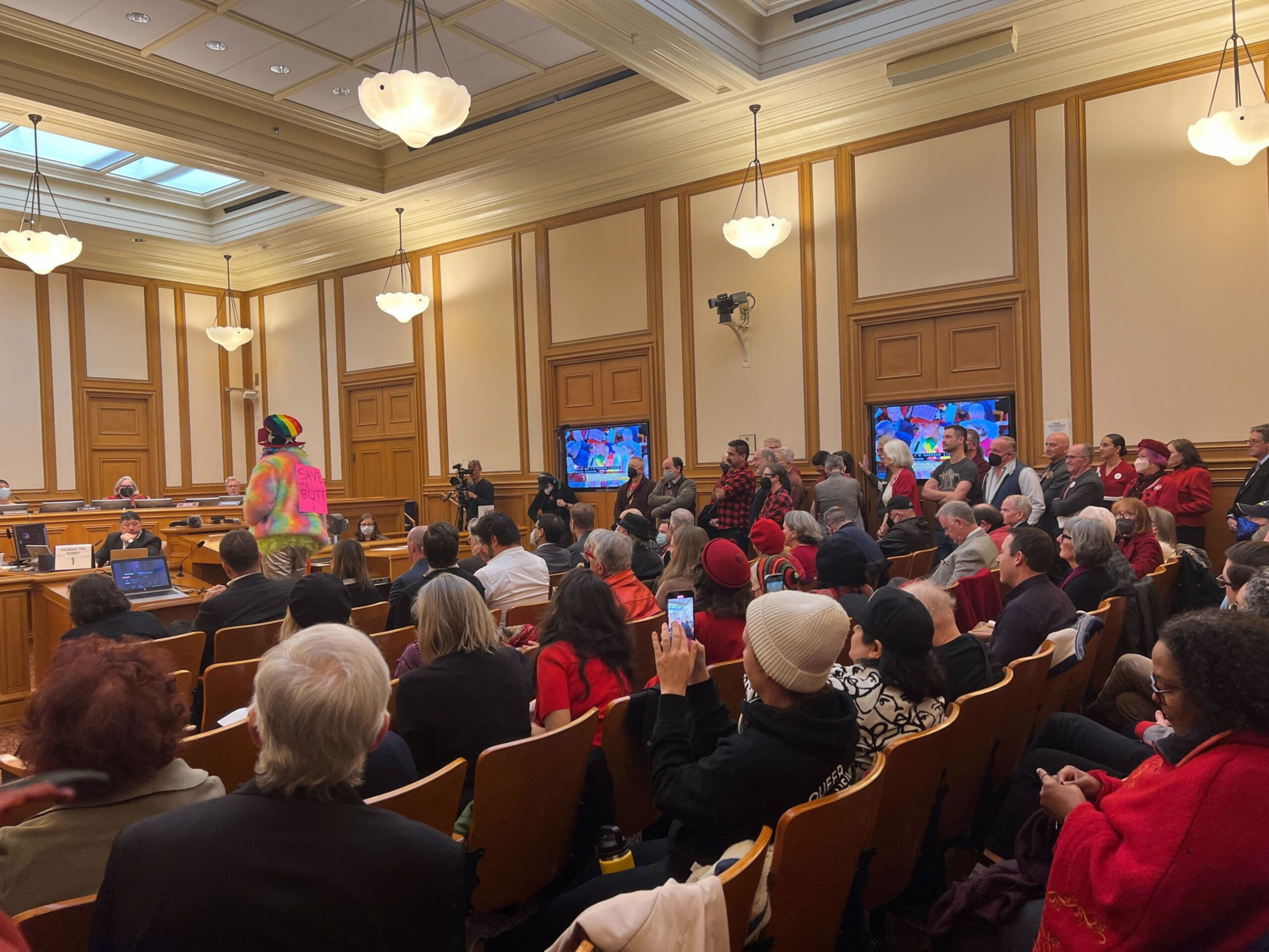 A person in a colorful hoodie stands speaking in a wood-paneled room filled with seated and standing people, some recording the event on phones.