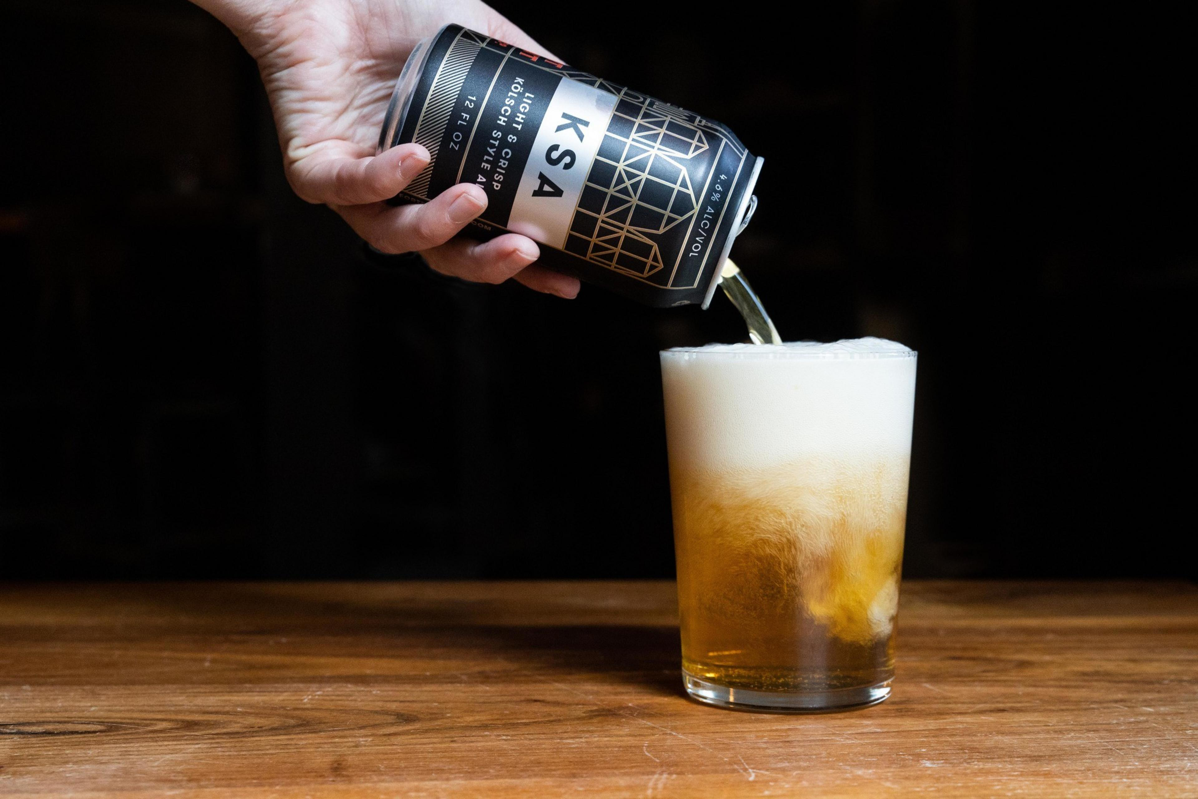 A hand pours a dark, geometric-patterned can of beer into a clear glass filled with frothy foam, set on a wooden surface against a dark background.