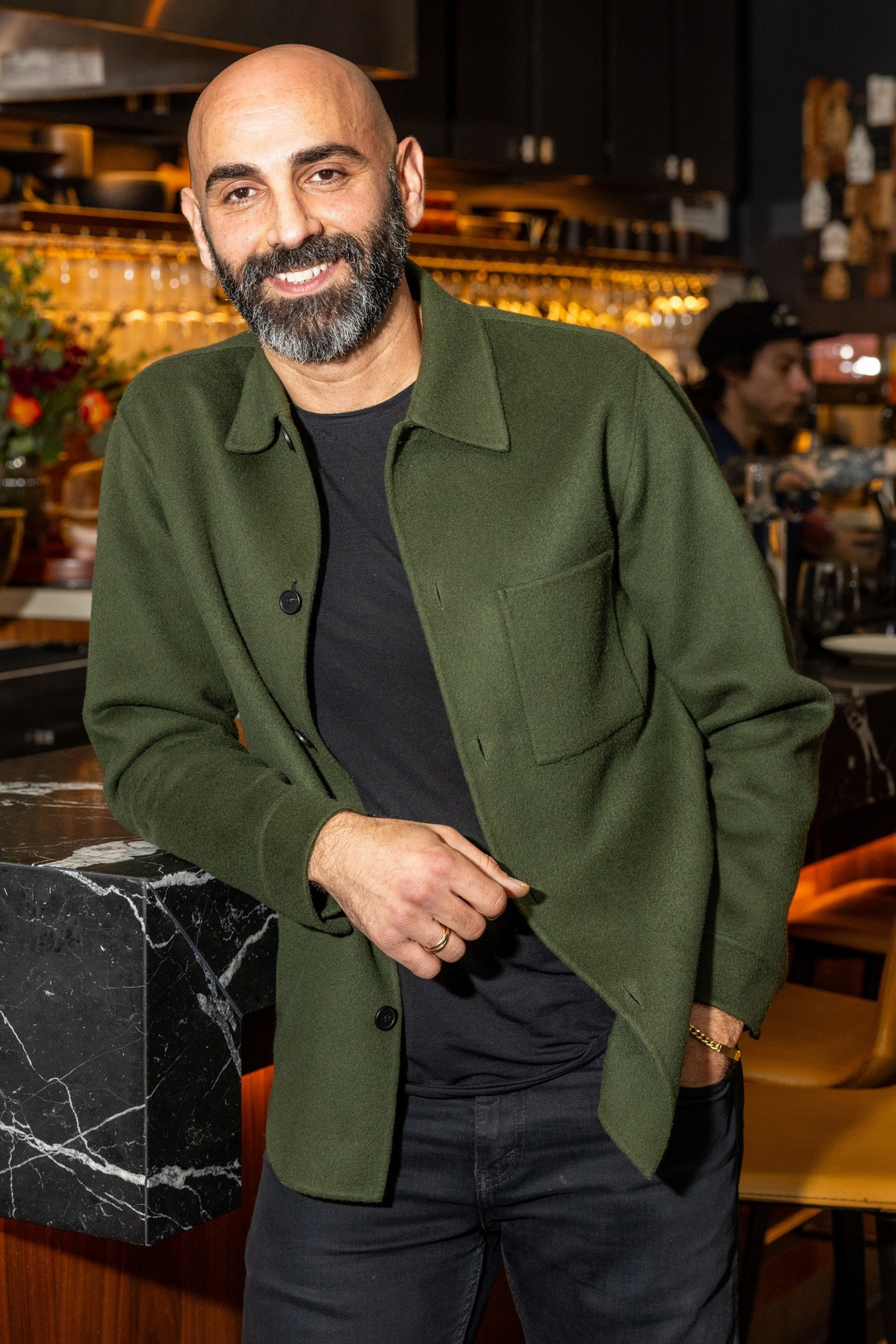 Ένας χαμογελαστός φαλακρός άνδρας με γένια φοράει ένα πράσινο σακάκι και μαύρο πουκάμισο, ακουμπισμένος χαλαρά σε έναν μαύρο μαρμάρινο πάγκο μπαρ σε ένα ζεστό φωτισμένο εστιατόριο.