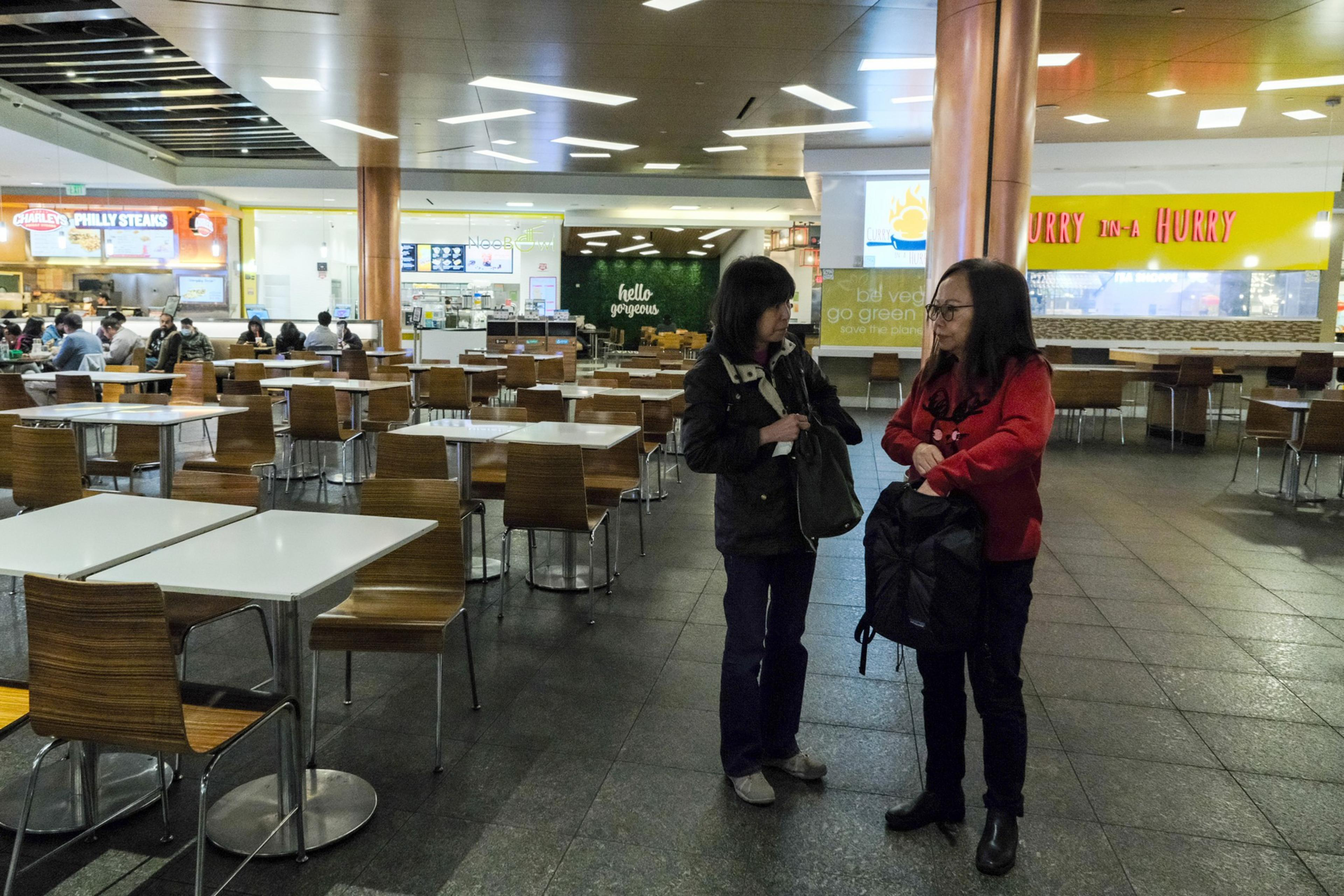 Two women talk in a mall food court with tables, chairs, and fast-food outlets in the background.