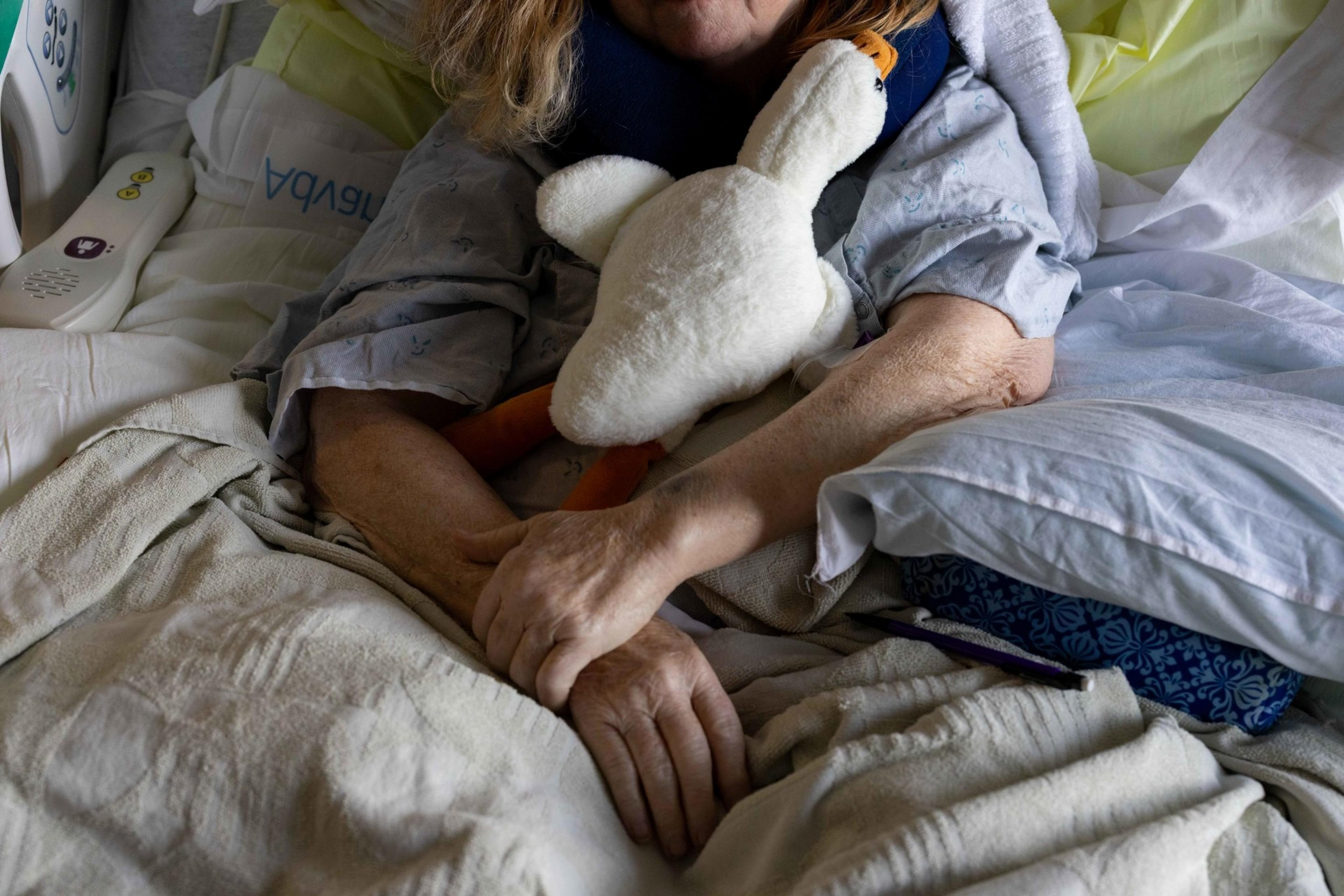 A person lies in a hospital bed, holding a stuffed animal. Their hands are clasped, with a blue neck pillow visible. Hospital sheets and a remote are nearby.
