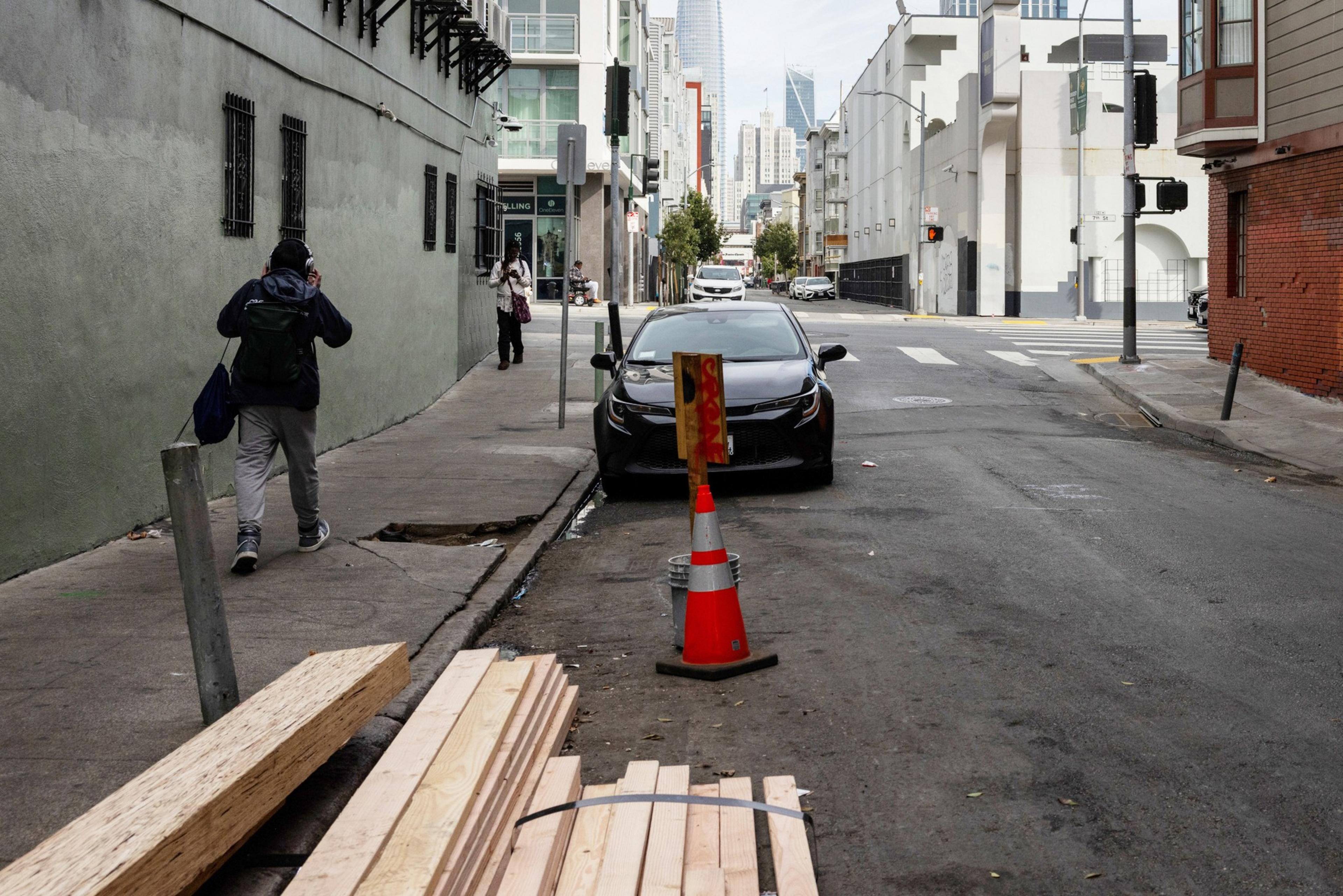 A pedestrian passes plywood laying on the street in a empty alleyway on a city block.