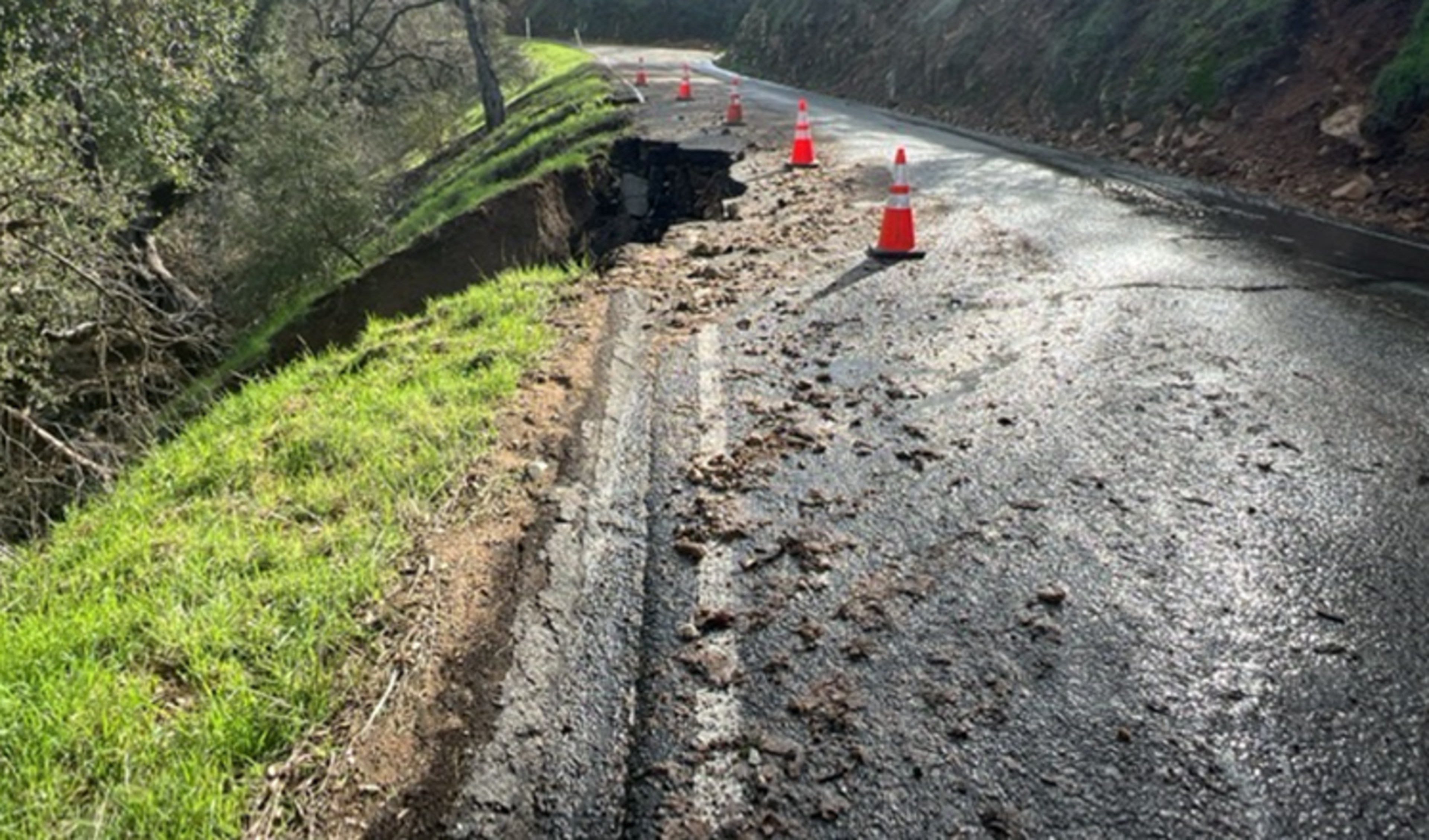 A rural road with a large sinkhole on the left side, marked by orange safety cones, and loose dirt scattered across the asphalt.