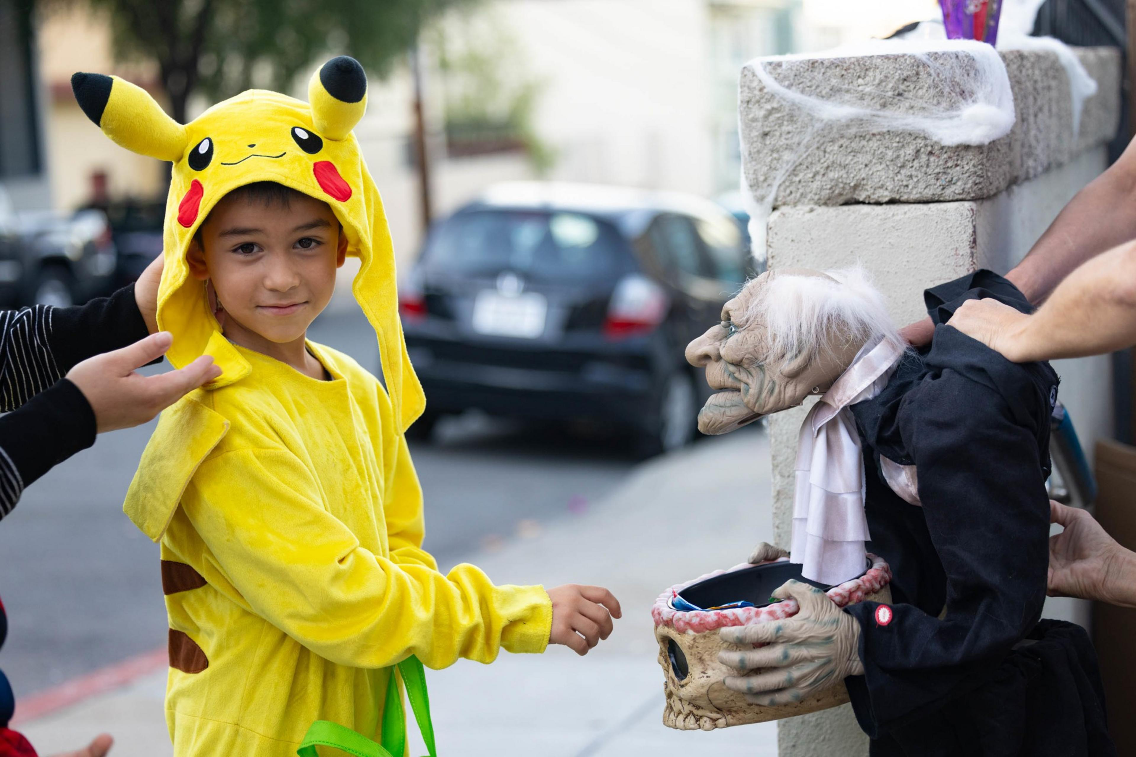 A child in a yellow Pikachu costume reaches toward a spooky puppet dressed in black, held by two adults near a stone wall on a street.