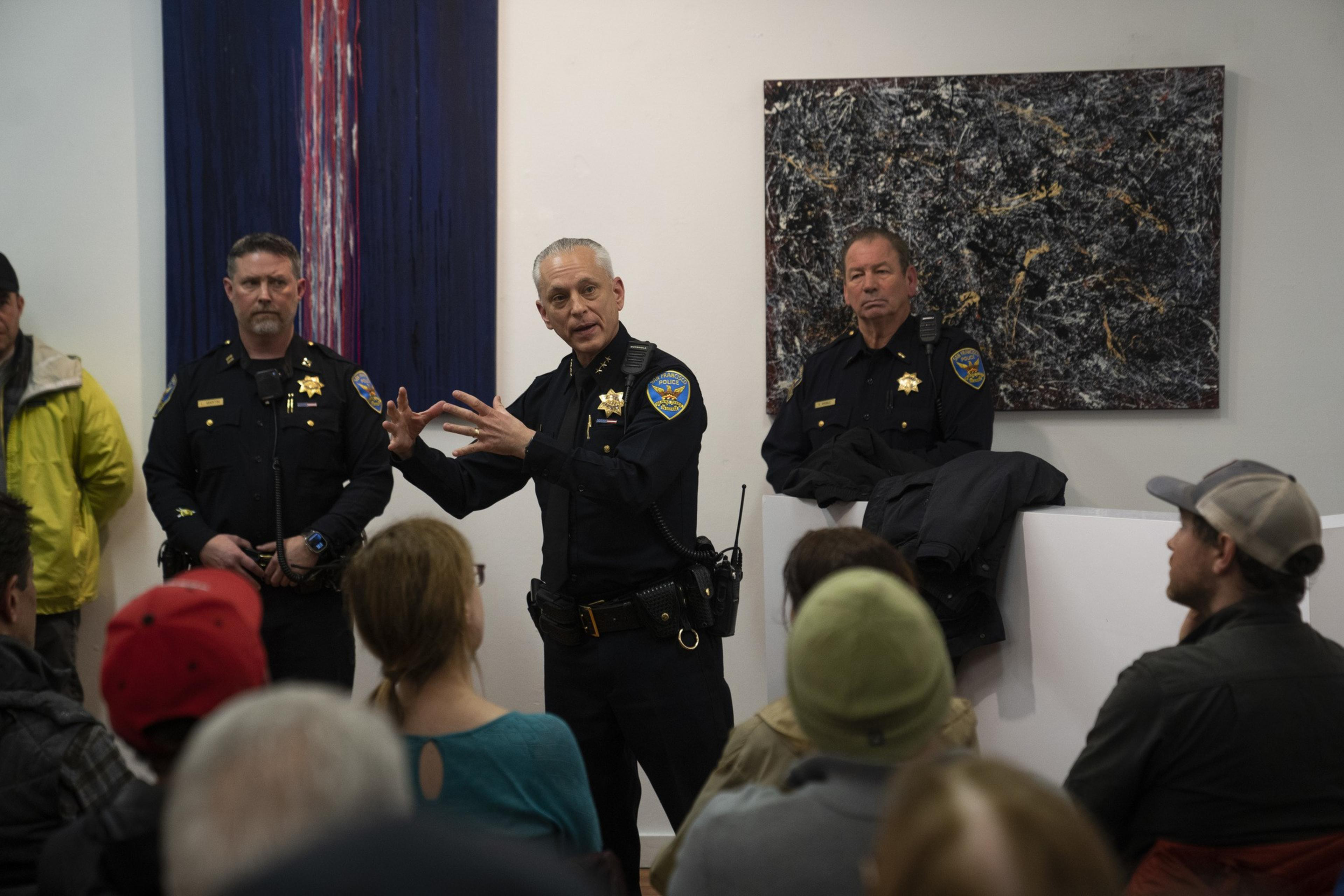 A police officer is speaking to a seated audience, gesturing with his hands. Two other officers stand nearby. Abstract paintings hang on the white walls behind them.