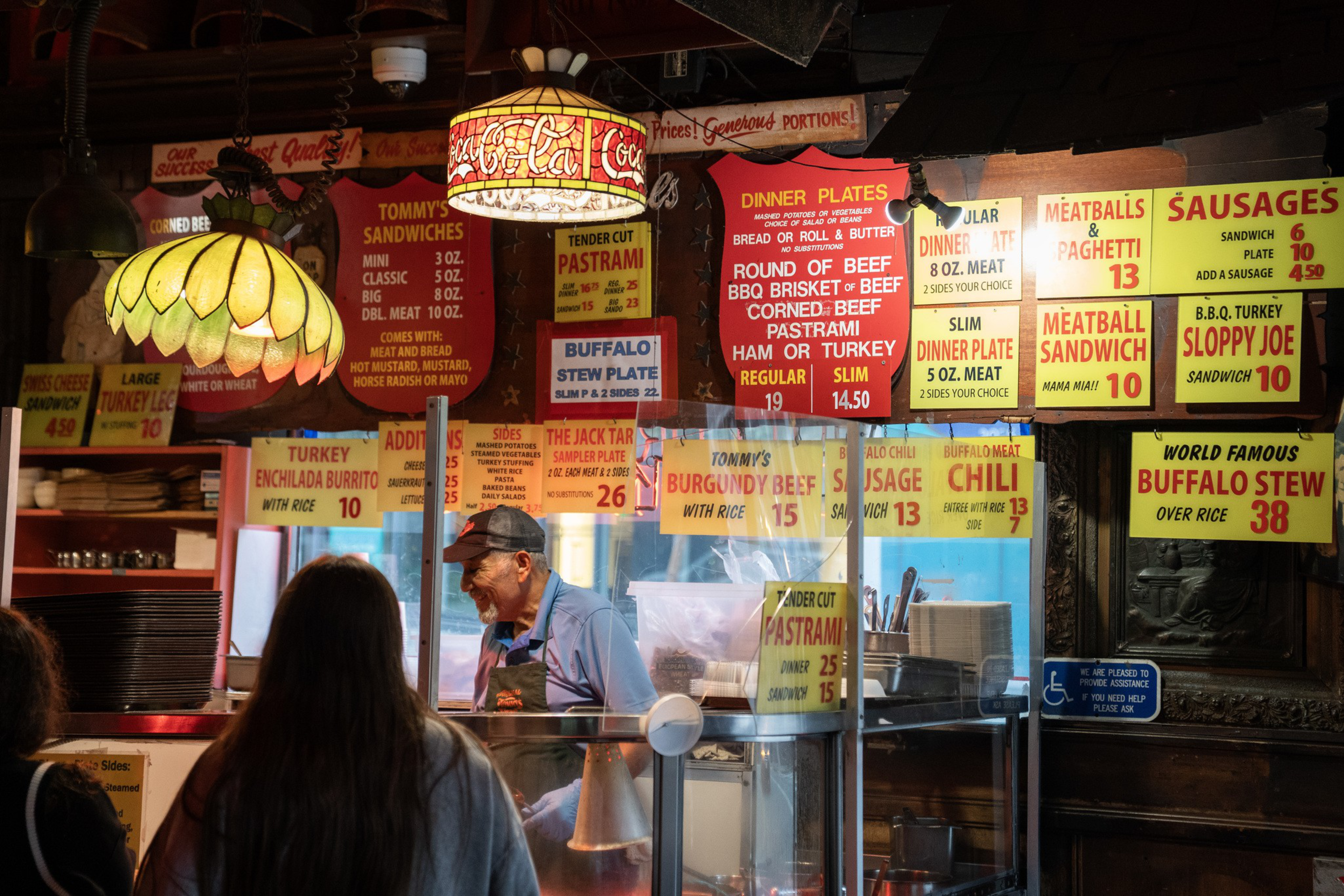 A deli counter with colorful hanging menus displays various sandwich, dinner plate, and specialty item options, including pastrami, meatballs, chili, and buffalo stew.