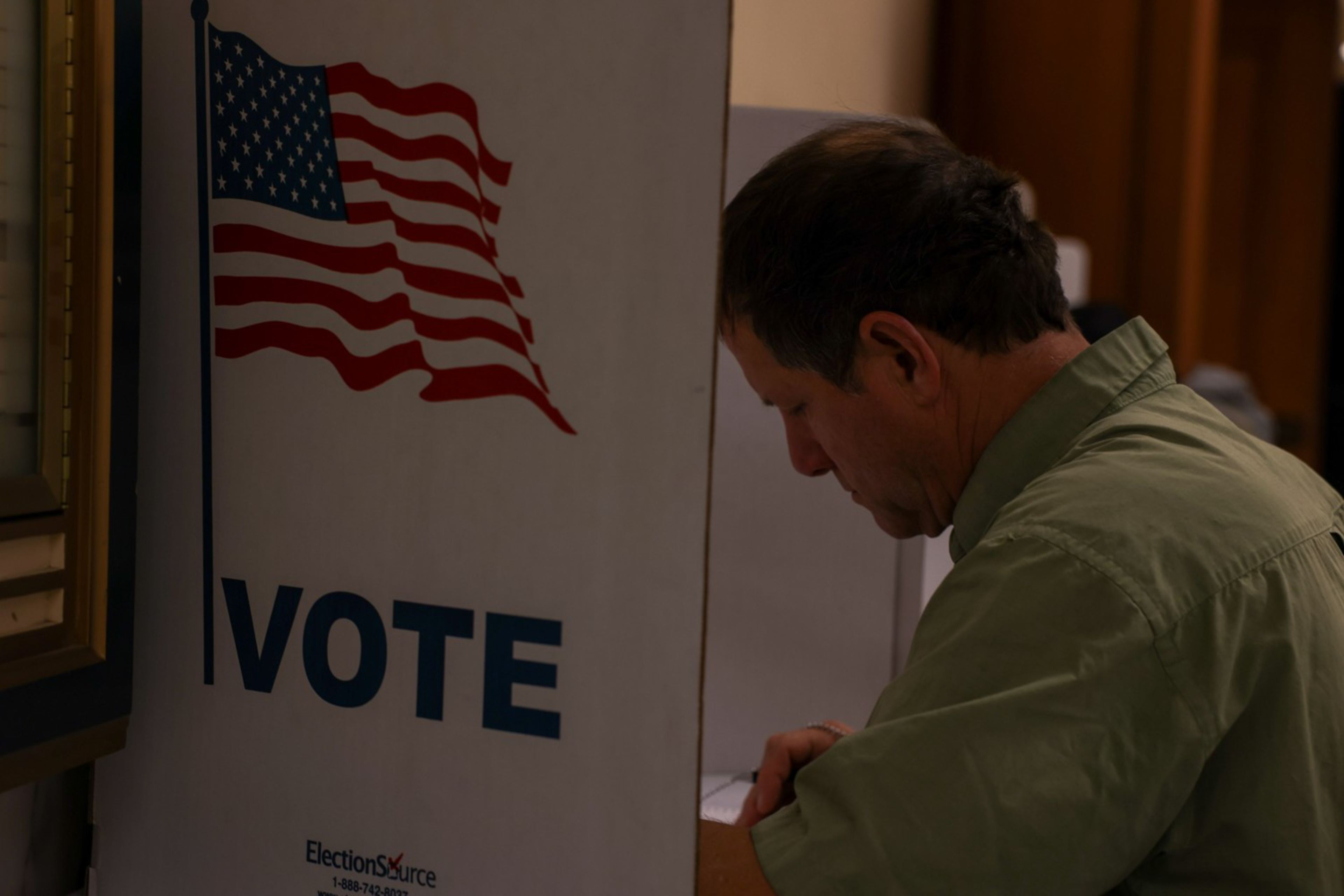 A man is filling out a ballot at a voting booth marked with an American flag and "VOTE."
