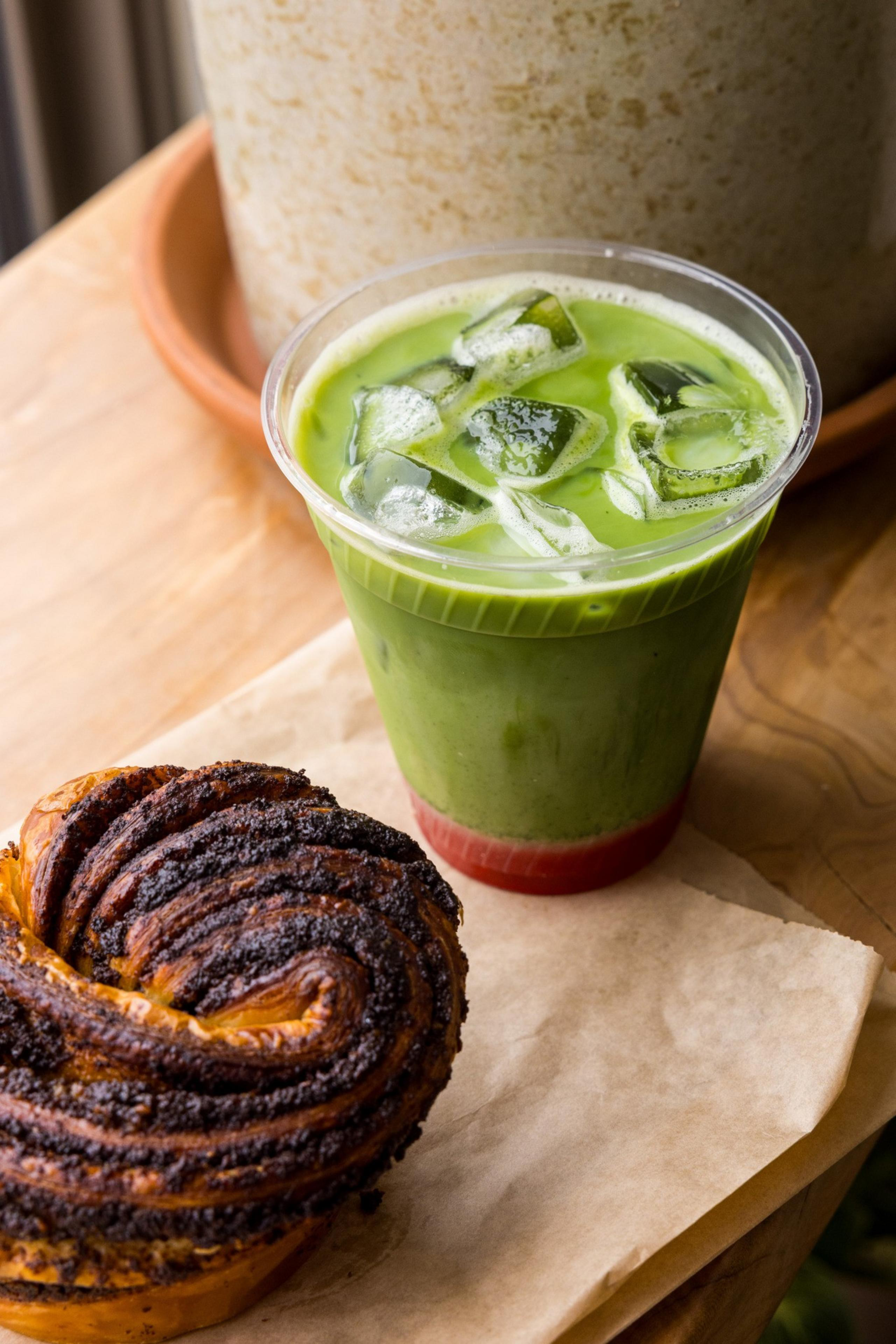 A swirl pastry sits on parchment paper next to a cup of iced green drink on a wooden table. The drink has ice cubes and a layered appearance with red at the bottom.