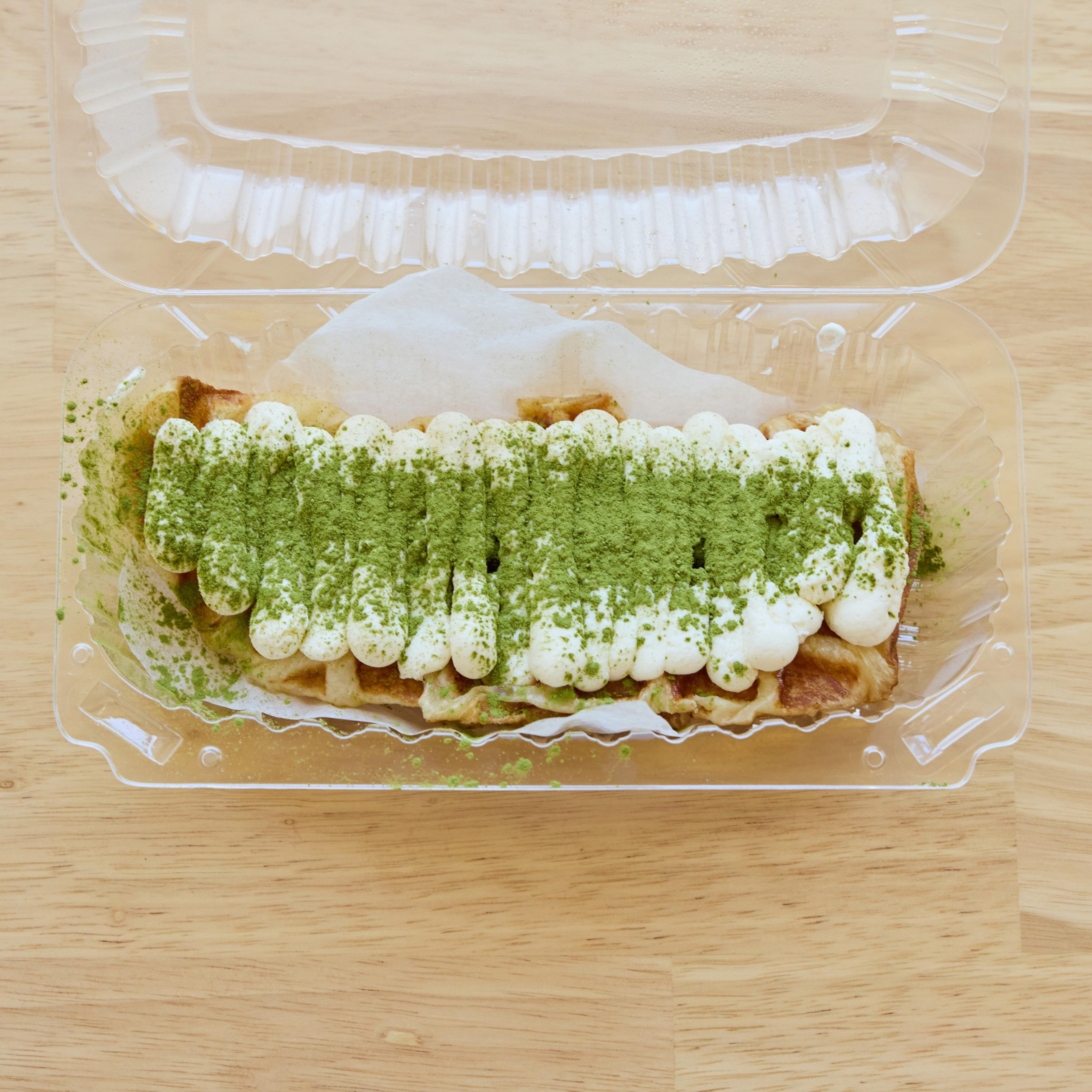 This image shows a waffle topped with whipped cream and a dusting of green matcha powder, inside a clear plastic takeout container.