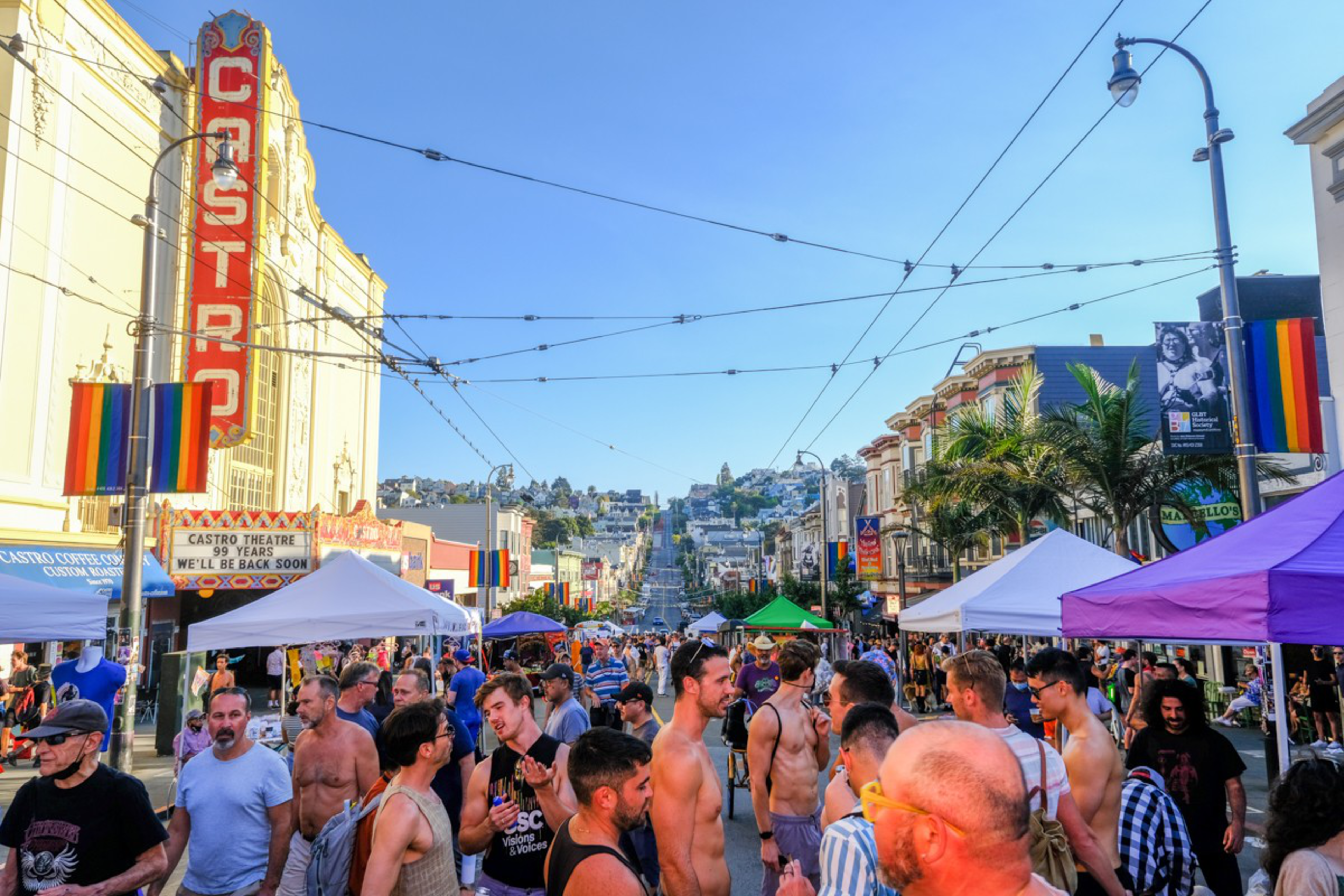 Crowds gather on a bright street lined with colorful flags and tents, near a landmark theater with a large vertical sign. It’s a festive, busy scene.
