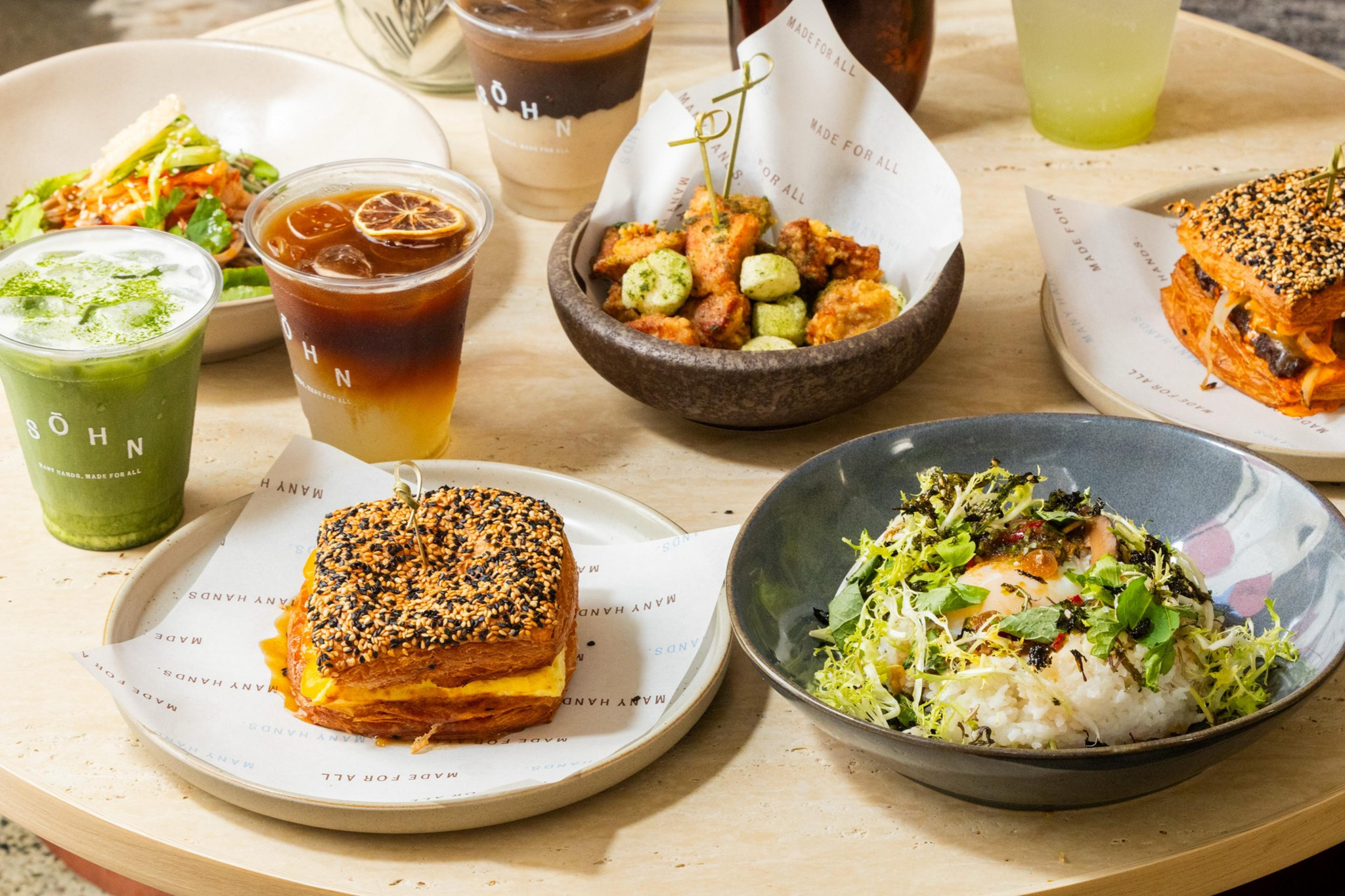 Two sesame seed-topped sandwiches, a bowl of mixed fried bites, a green salad with shredded vegetables, a rice bowl with greens and egg, and three iced drinks.