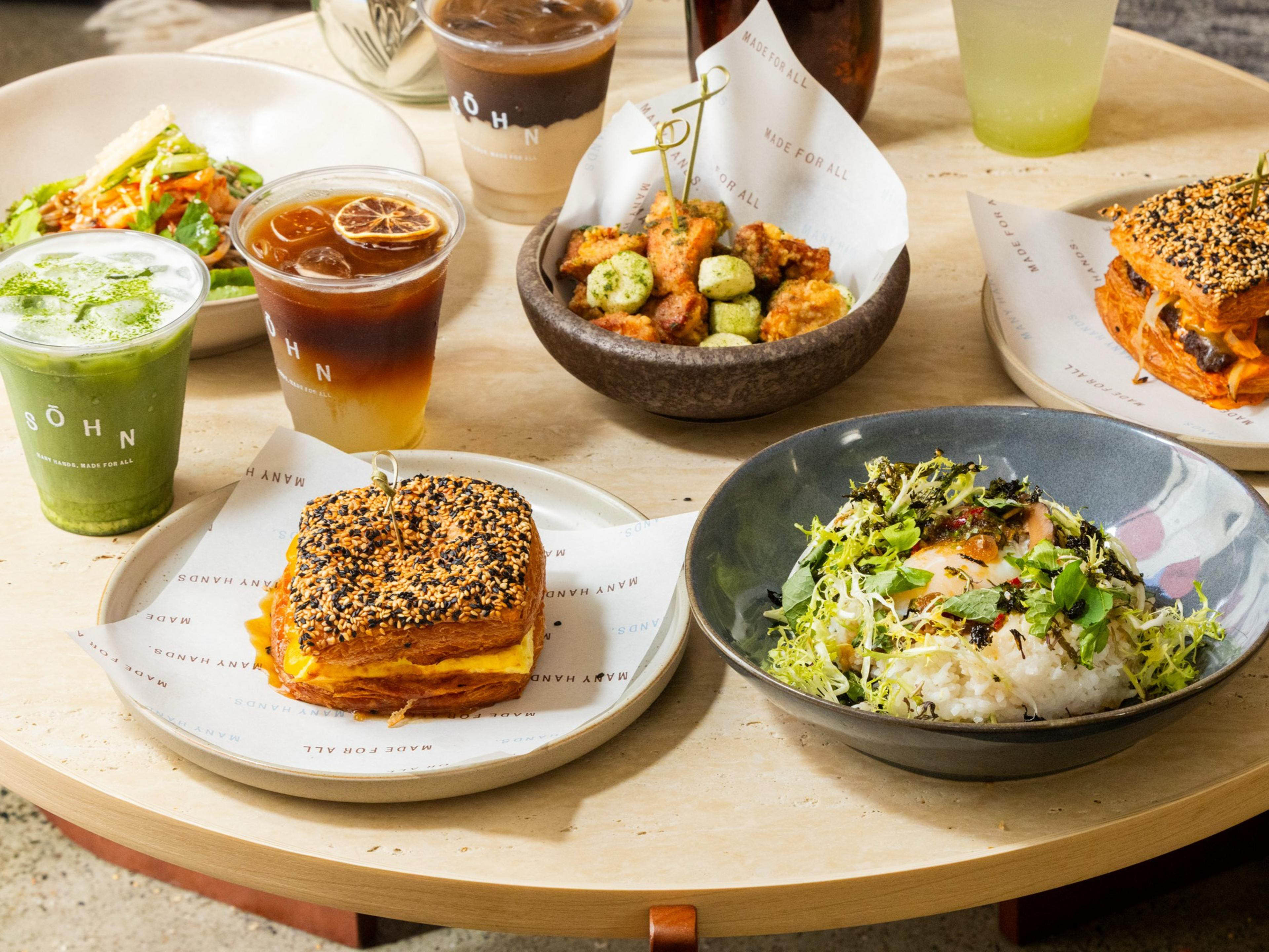 Two sesame seed-topped sandwiches, a bowl of mixed fried bites, a green salad with shredded vegetables, a rice bowl with greens and egg, and three iced drinks.