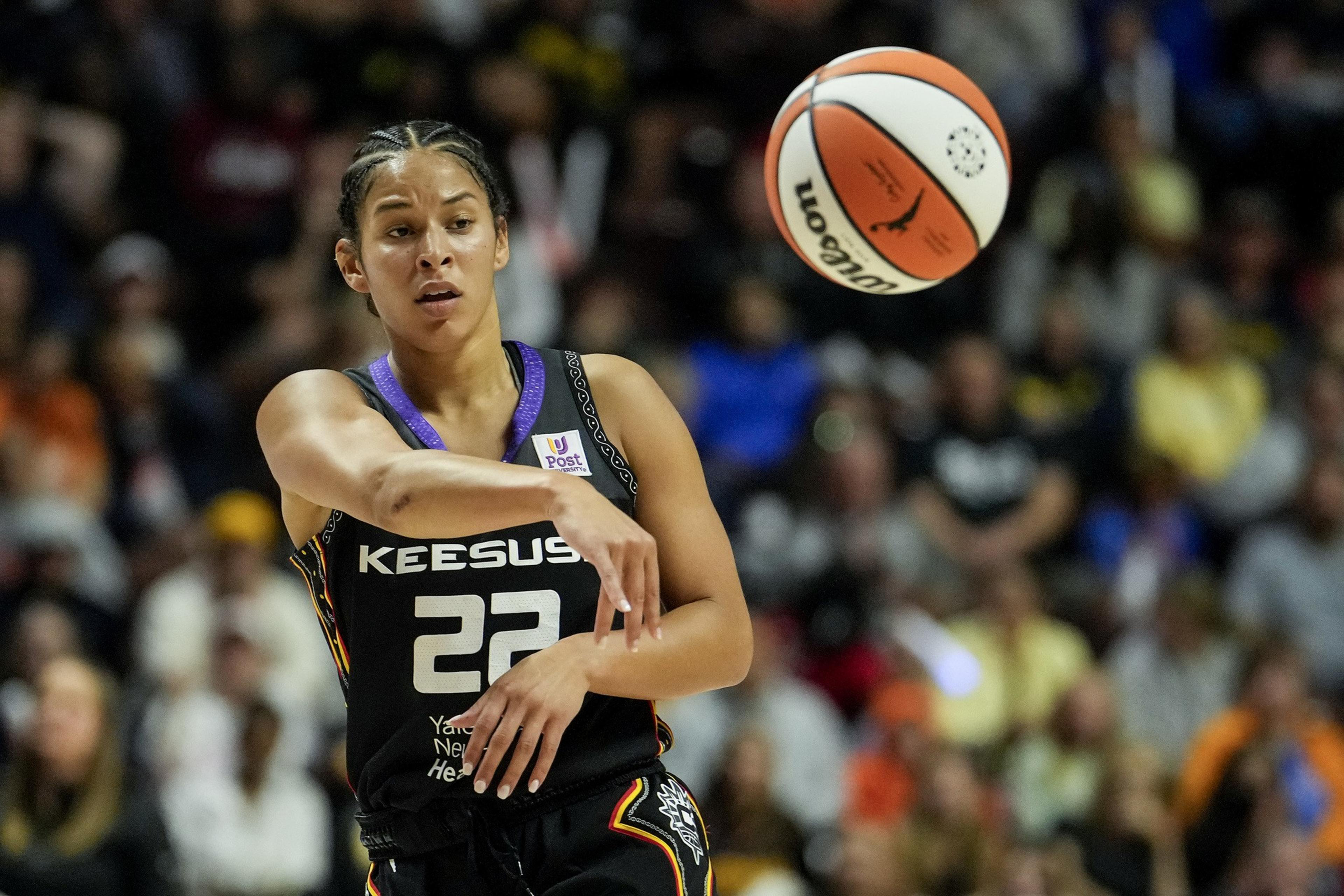 A basketball player in a black jersey with number 22 is focused as she throws a basketball. The crowd in the background is blurred.