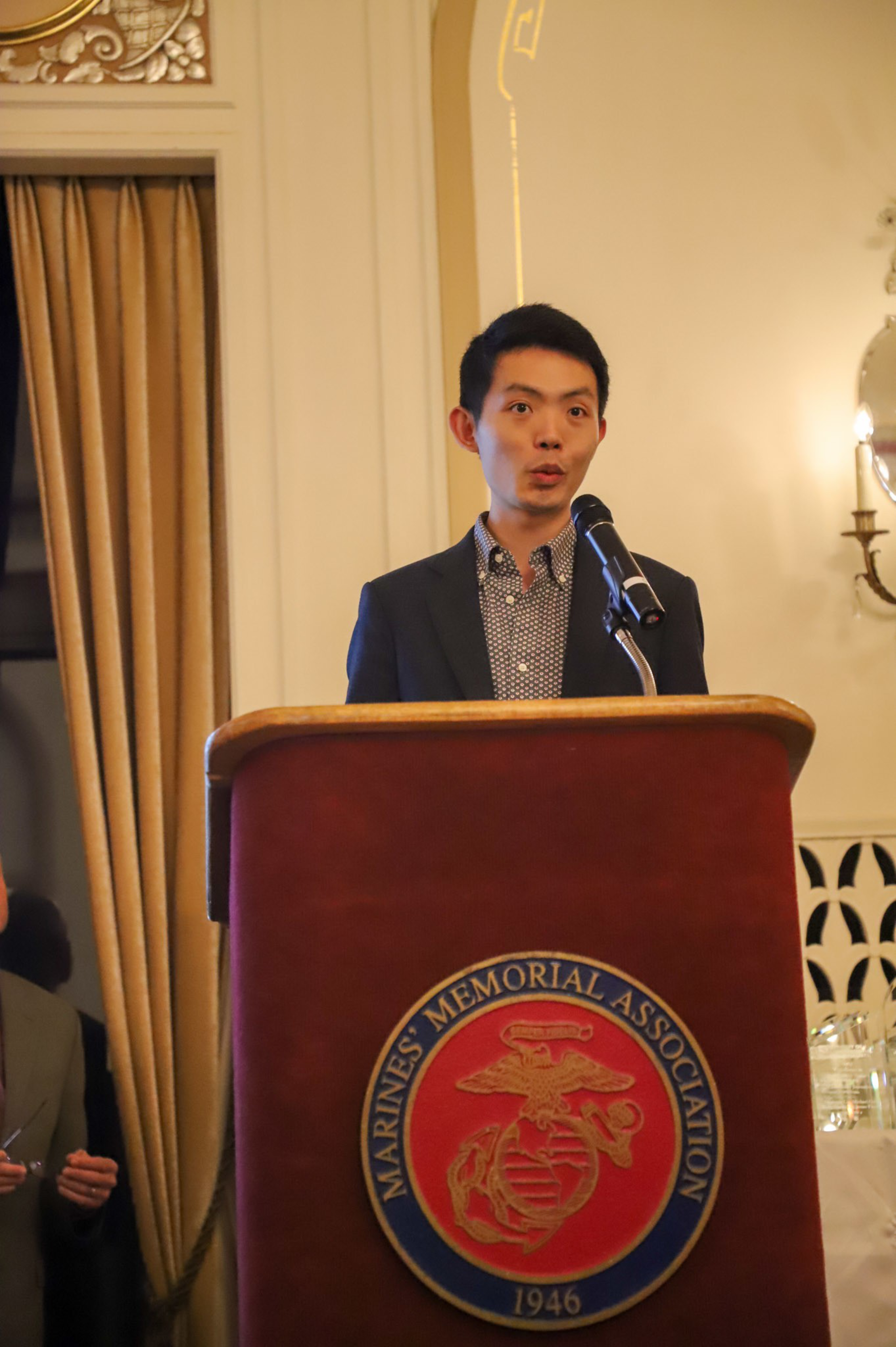 A man in a suit stands behind a podium with a Marines' Memorial Association emblem, speaking into a microphone in a formal indoor setting.