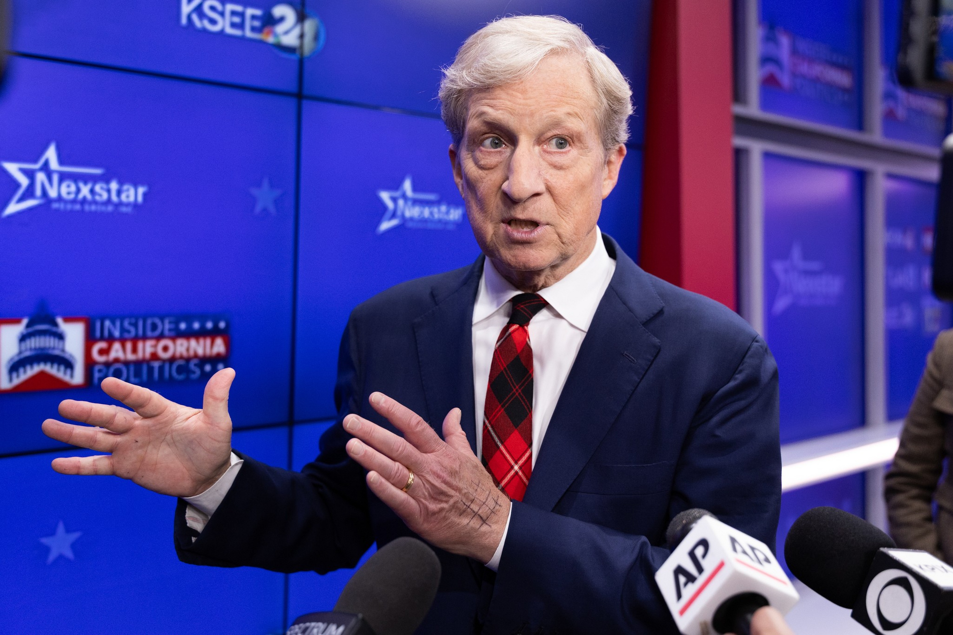 A man with gray hair in a dark suit and red plaid tie speaks and gestures in front of microphones labeled AP and CBS at a political event.