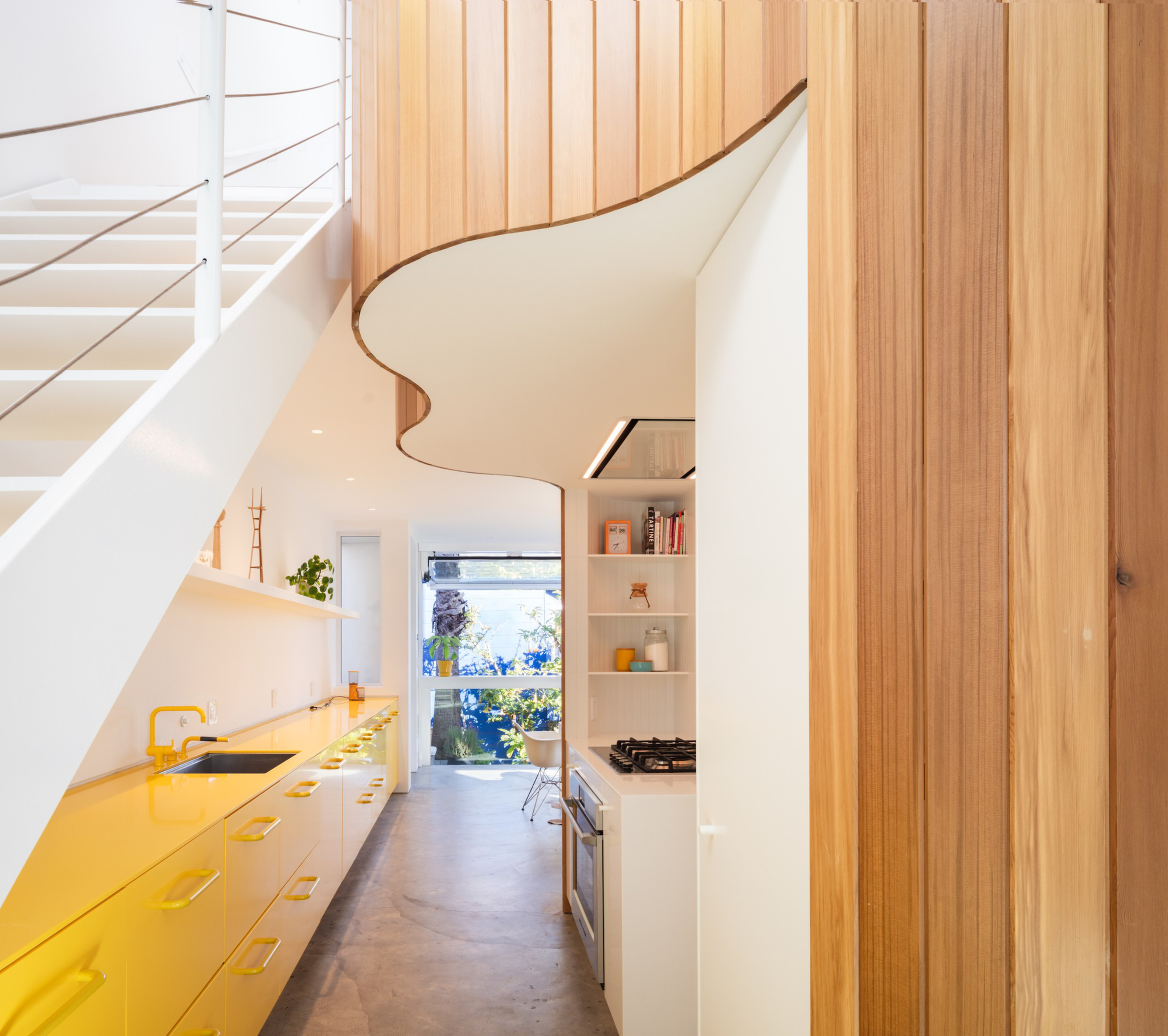 A modern kitchen with yellow countertops, white cabinets, and wooden paneling. A staircase with white rails is visible on the left. Natural light floods in.