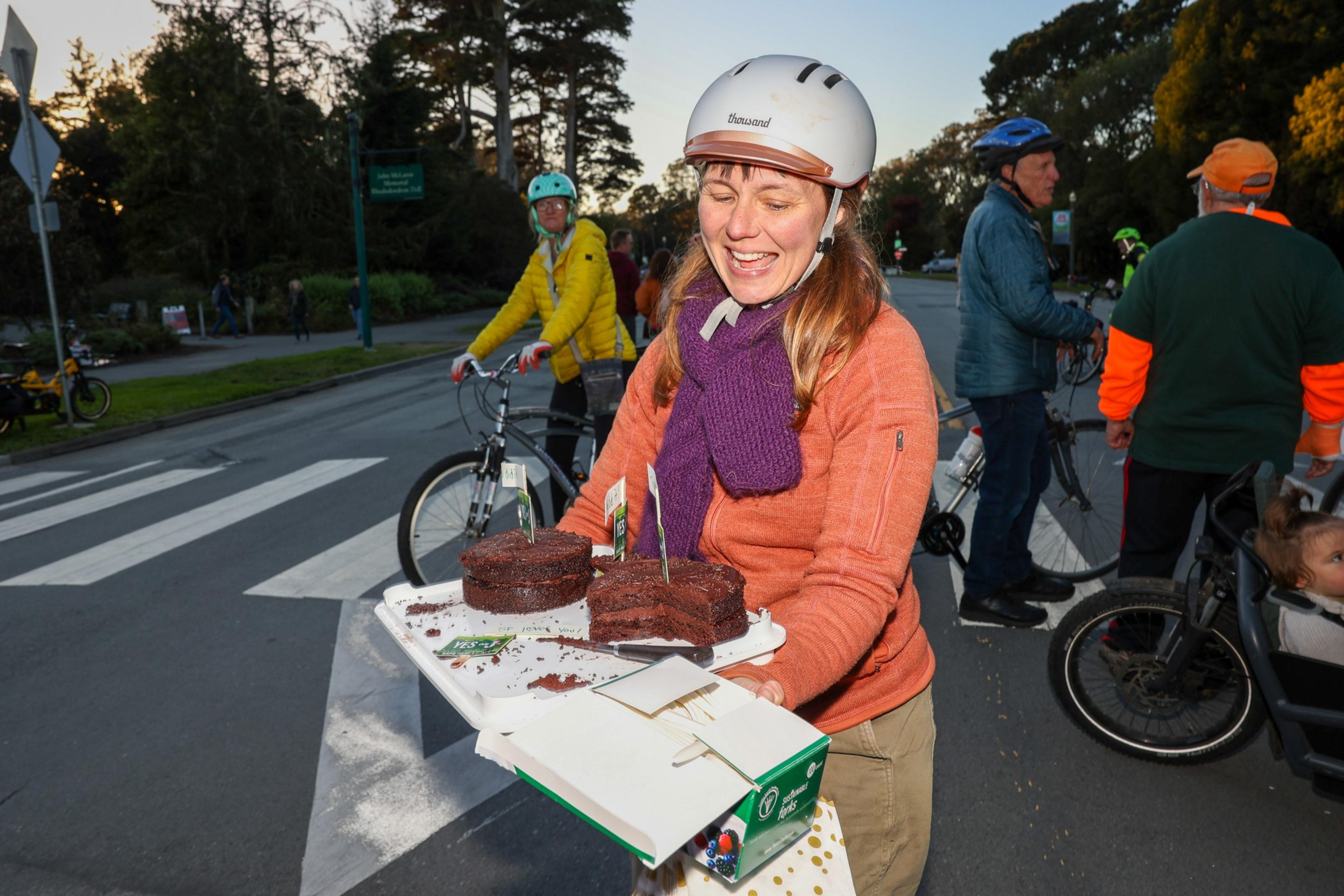 A woman wearing a white helmet and purple scarf holds a partially eaten chocolate cake outdoors, with cyclists and trees in the background.