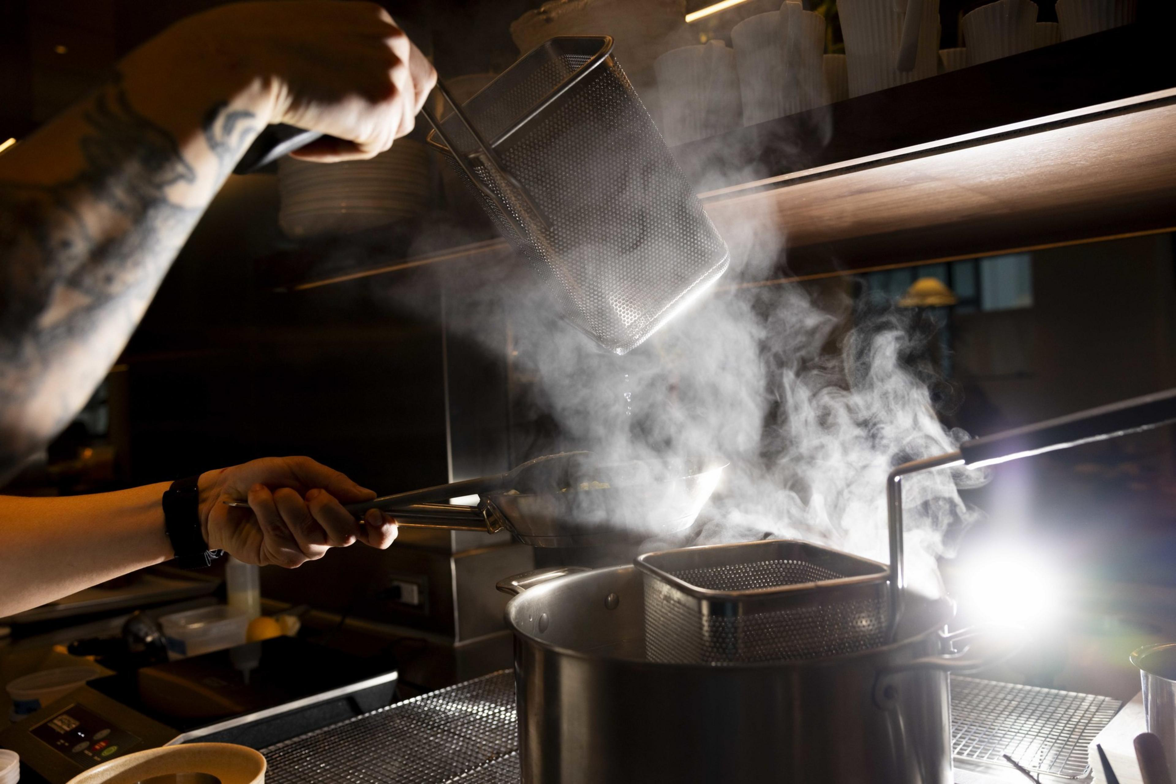 A person is cooking, holding a slotted spoon and a sieve over a steaming pot. Their tattooed arm is visible, and the kitchen is dimly lit.