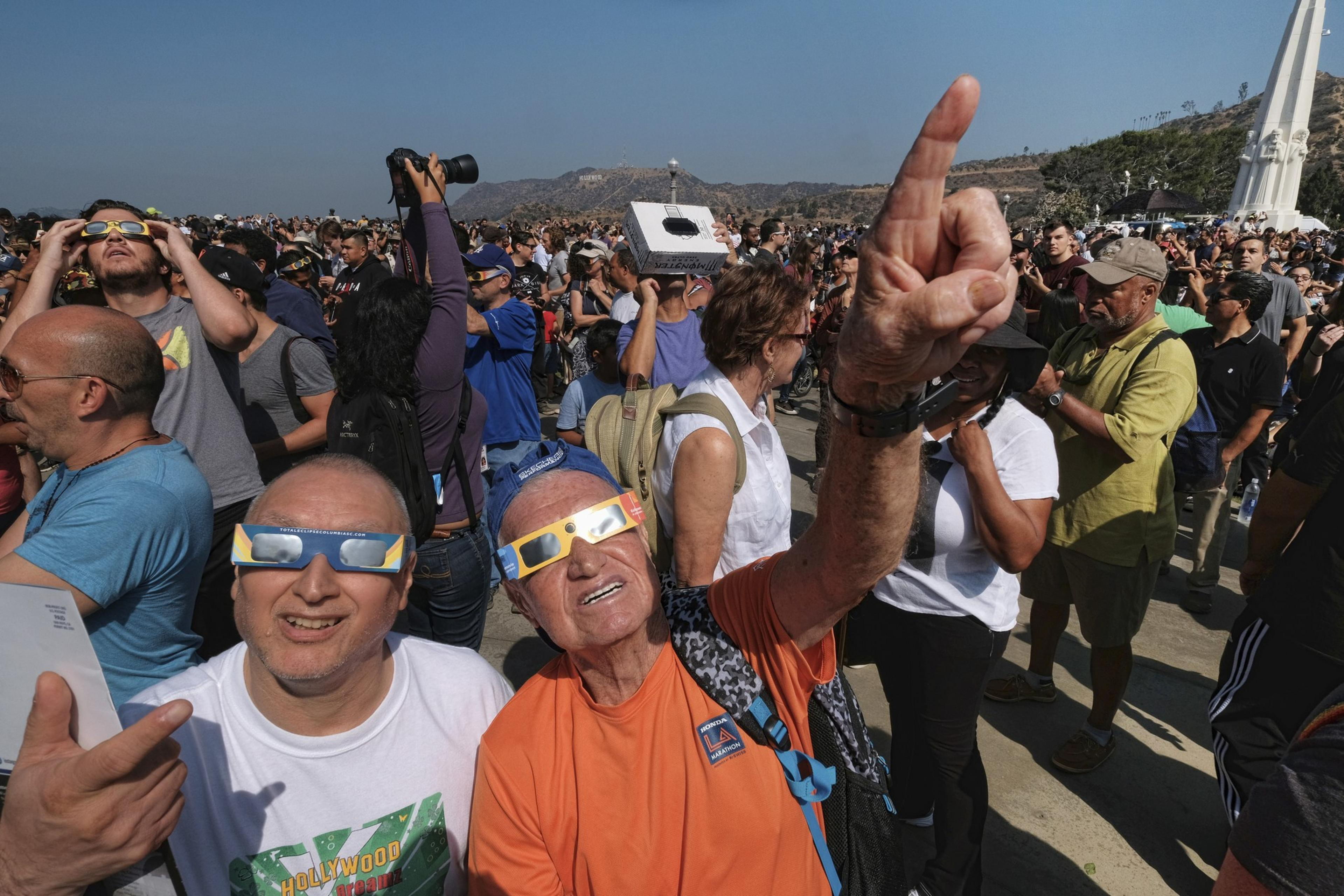 A crowd wearing special glasses looks up at the sky, likely viewing an eclipse, with excited expressions and gestures.