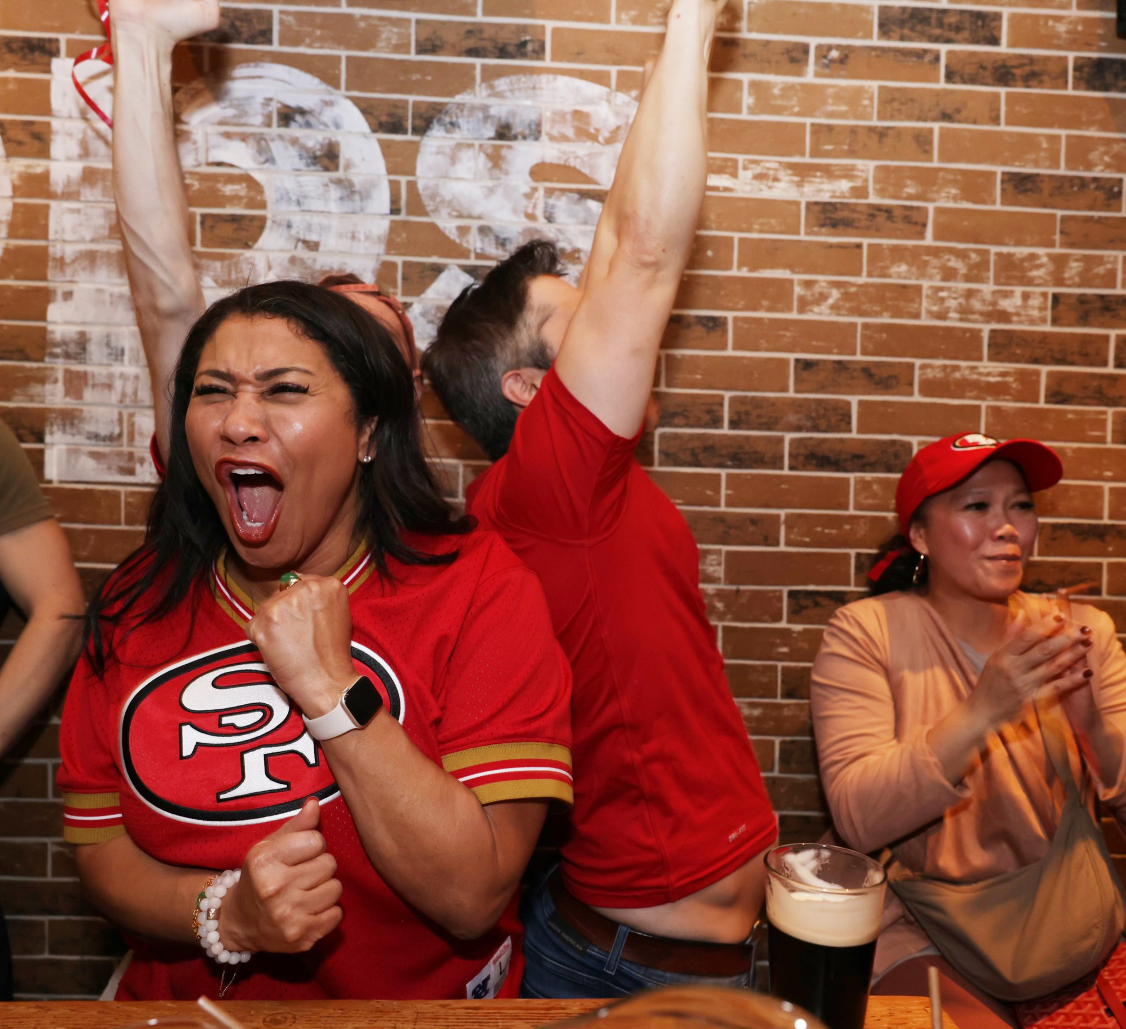 Three people celebrate enthusiastically at a bar, wearing red sports apparel. A woman in front cheers with her fist raised, while the others clap and raise arms.