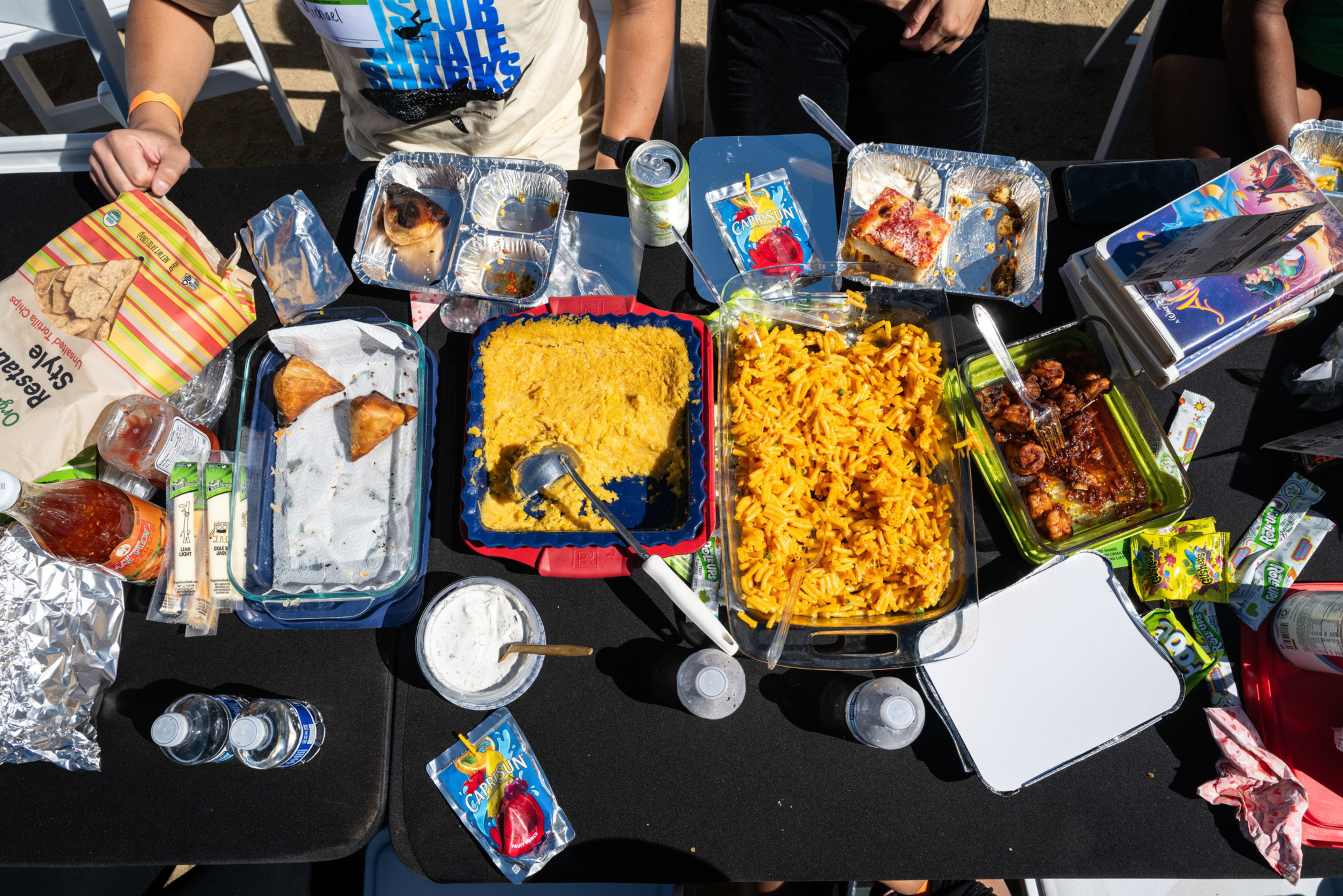 A table with various foods including samosas, macaroni and cheese, dip, barbecue chicken wings, juice pouches, water bottles, and snacks, with people seated around.