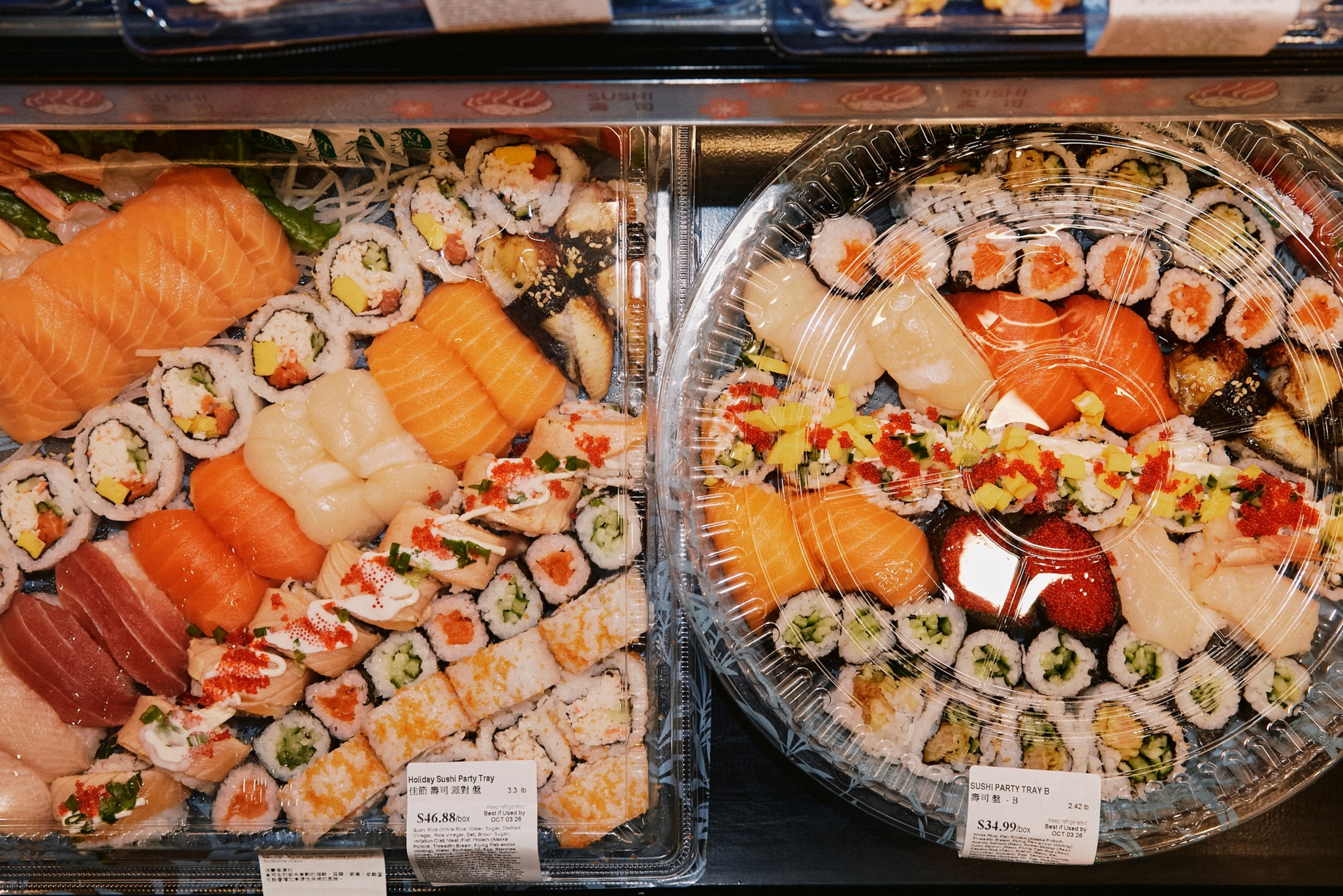 Two trays of assorted sushi, including salmon, tuna, rolls with avocado, cucumber, fish roe, and nigiri, are wrapped in clear plastic packaging.