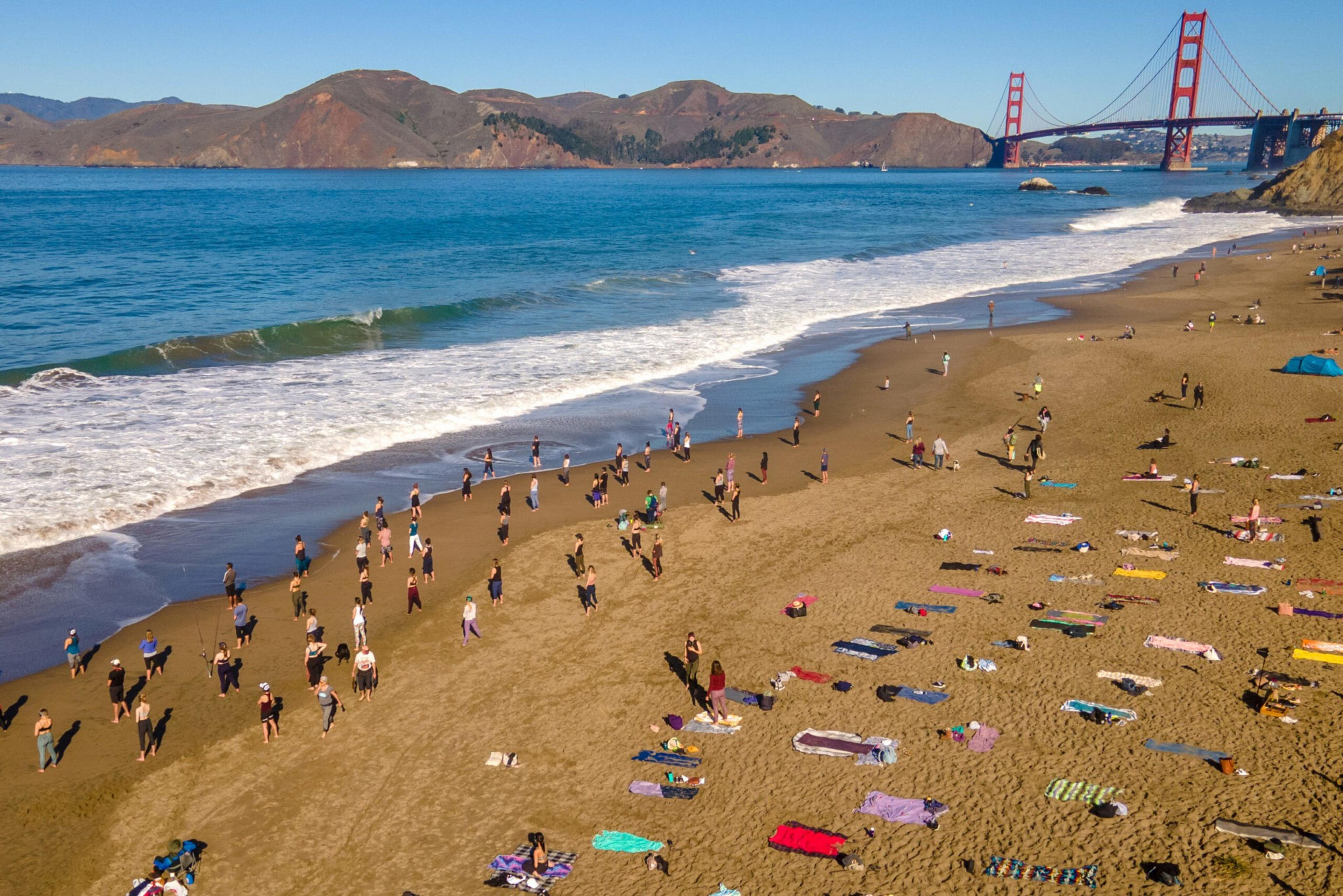 Many people practice yoga on a sandy beach near the ocean, with colorful towels spread around and the Golden Gate Bridge in the background.