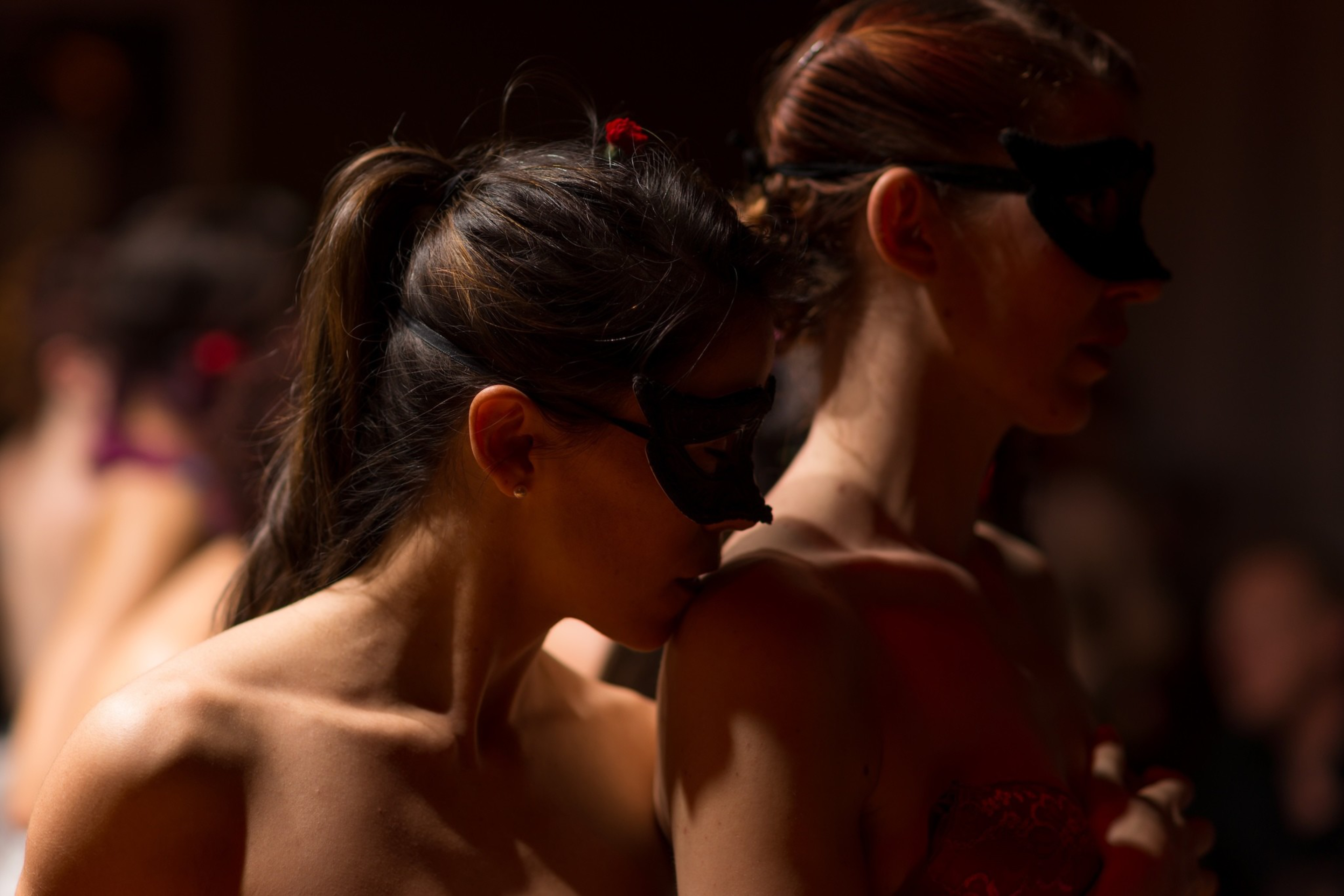 Two women wearing black masks stand closely, one gently touching the other’s shoulder with her lips in a dim, intimate setting.