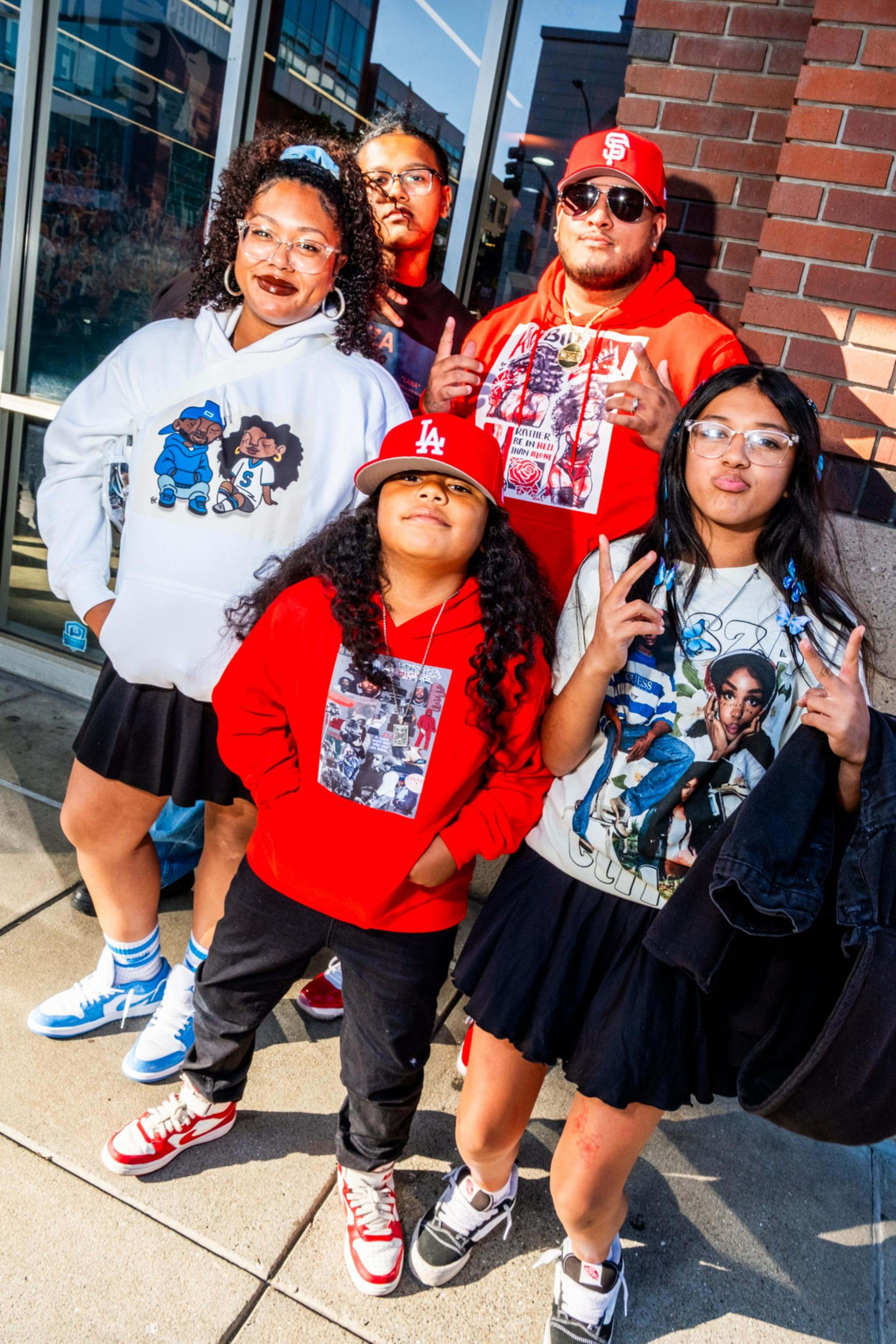 Five people pose in colorful outfits on a city sidewalk. They wear sneakers, hoodies, and graphic tees, with two making peace signs. They're smiling confidently.