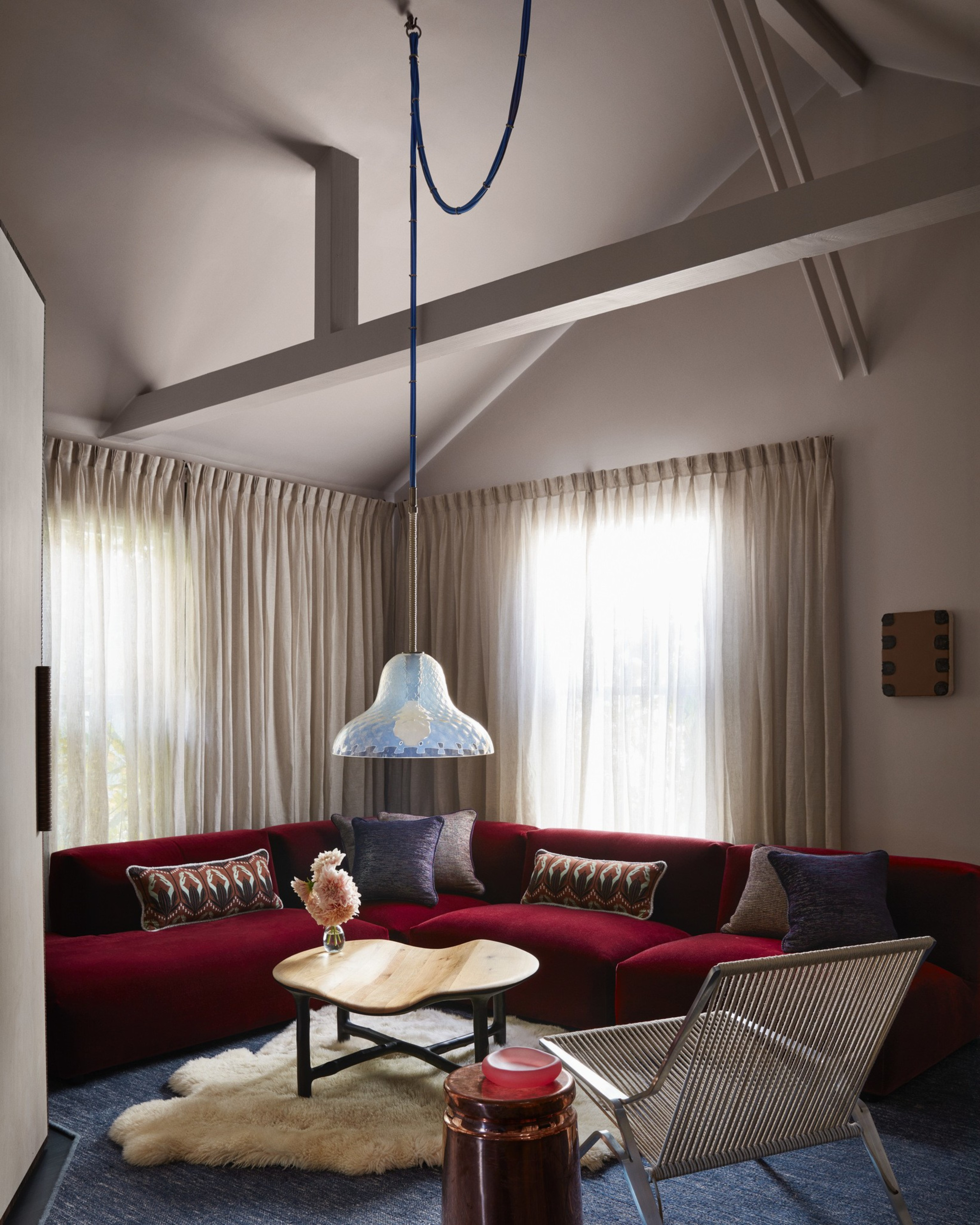 A cozy corner with a deep red L-shaped sofa, patterned pillows, a wooden coffee table, a furry rug, a metallic chair, and a hanging pendant light.
