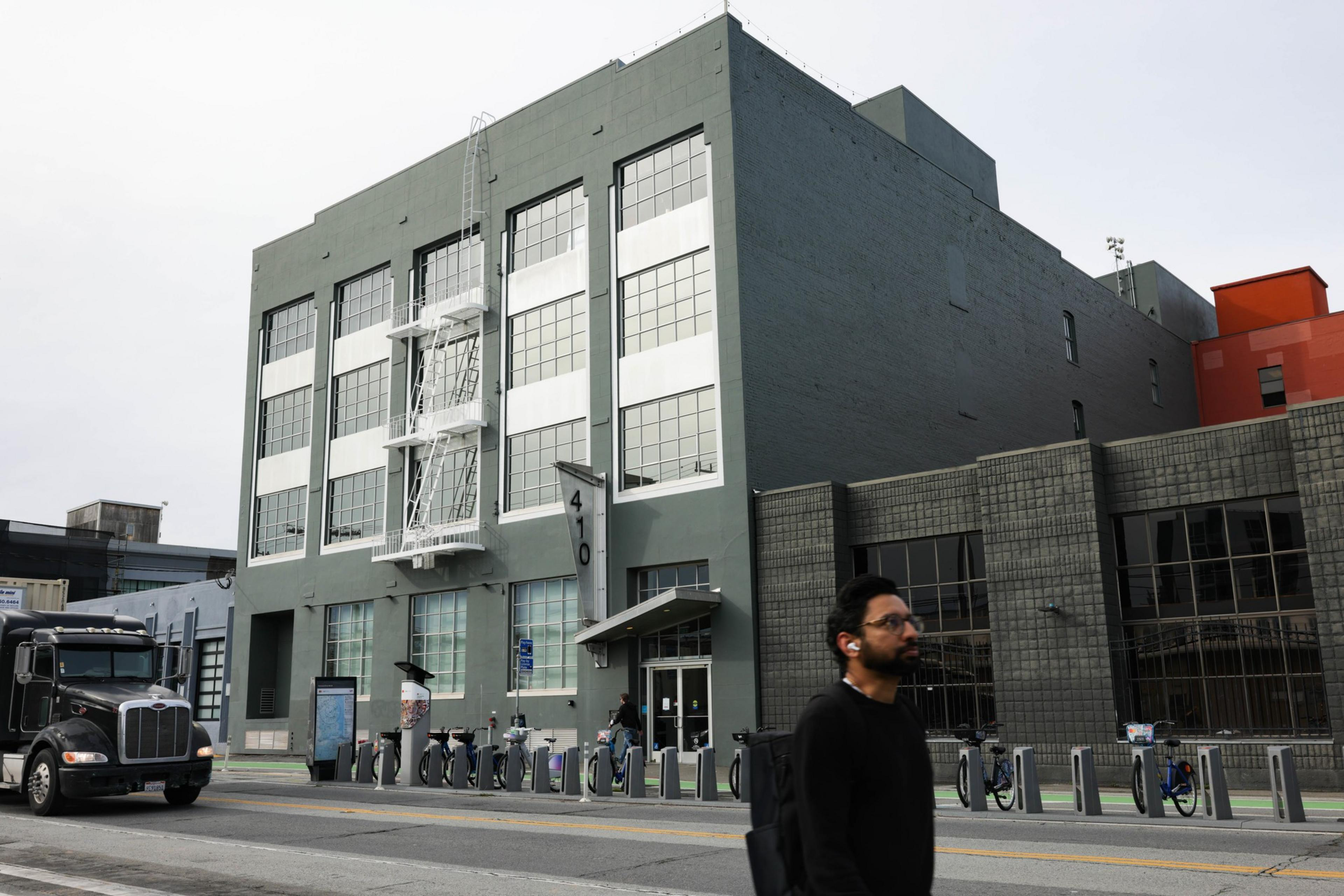 A person walks in front of a dark green building with a row of city bikes. 