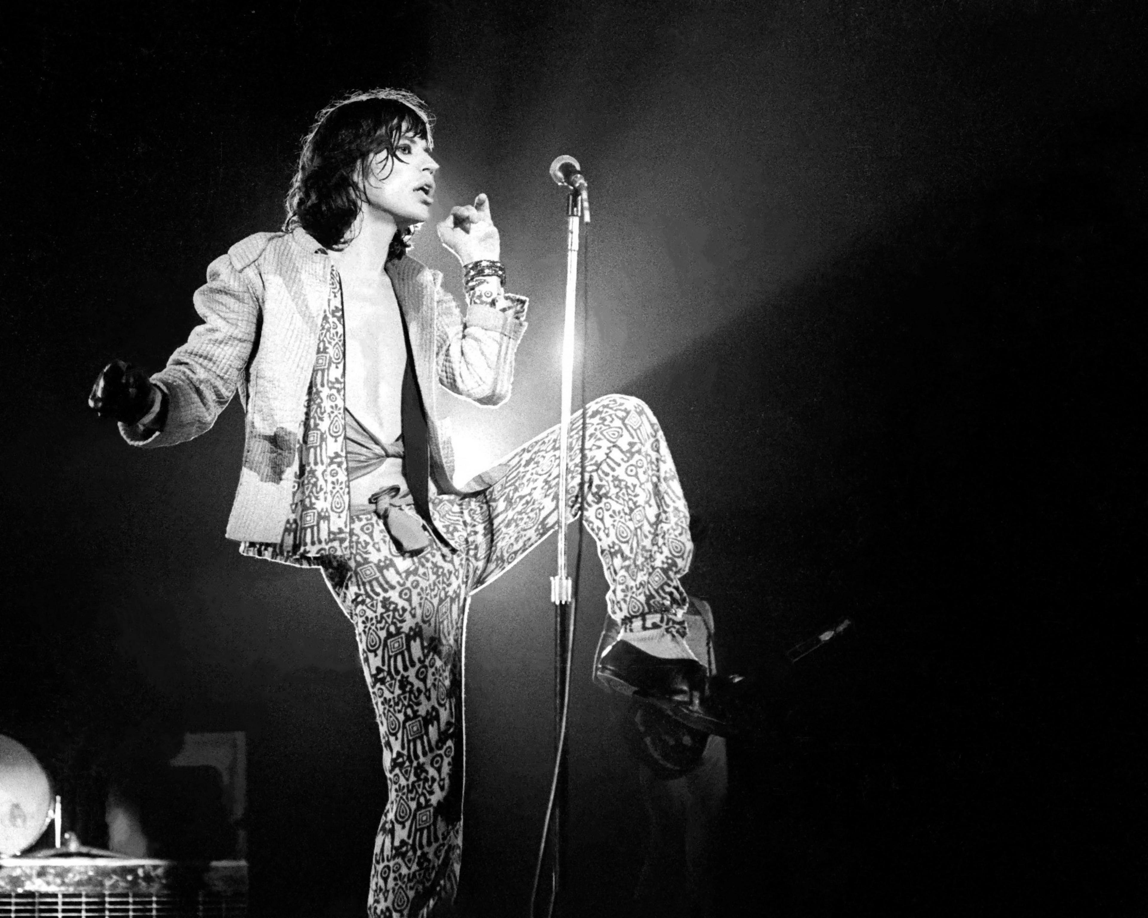 A performer in patterned pants and jacket sings at a microphone on stage, embodying a dynamic pose.