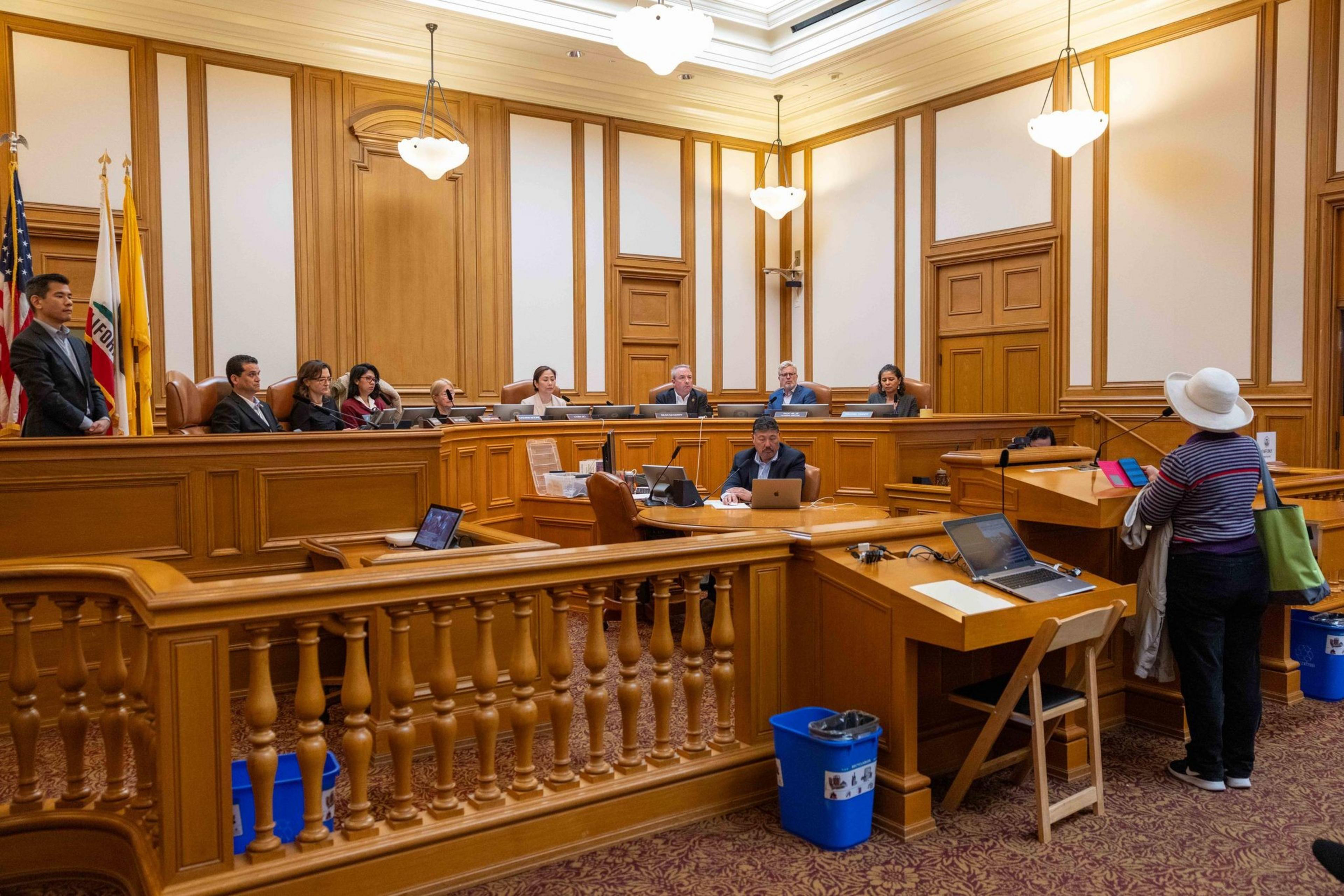 A person in a hat speaks at a podium in a wood-paneled room, addressing seated officials at a long desk. Laptops and flags add to the formal setting.