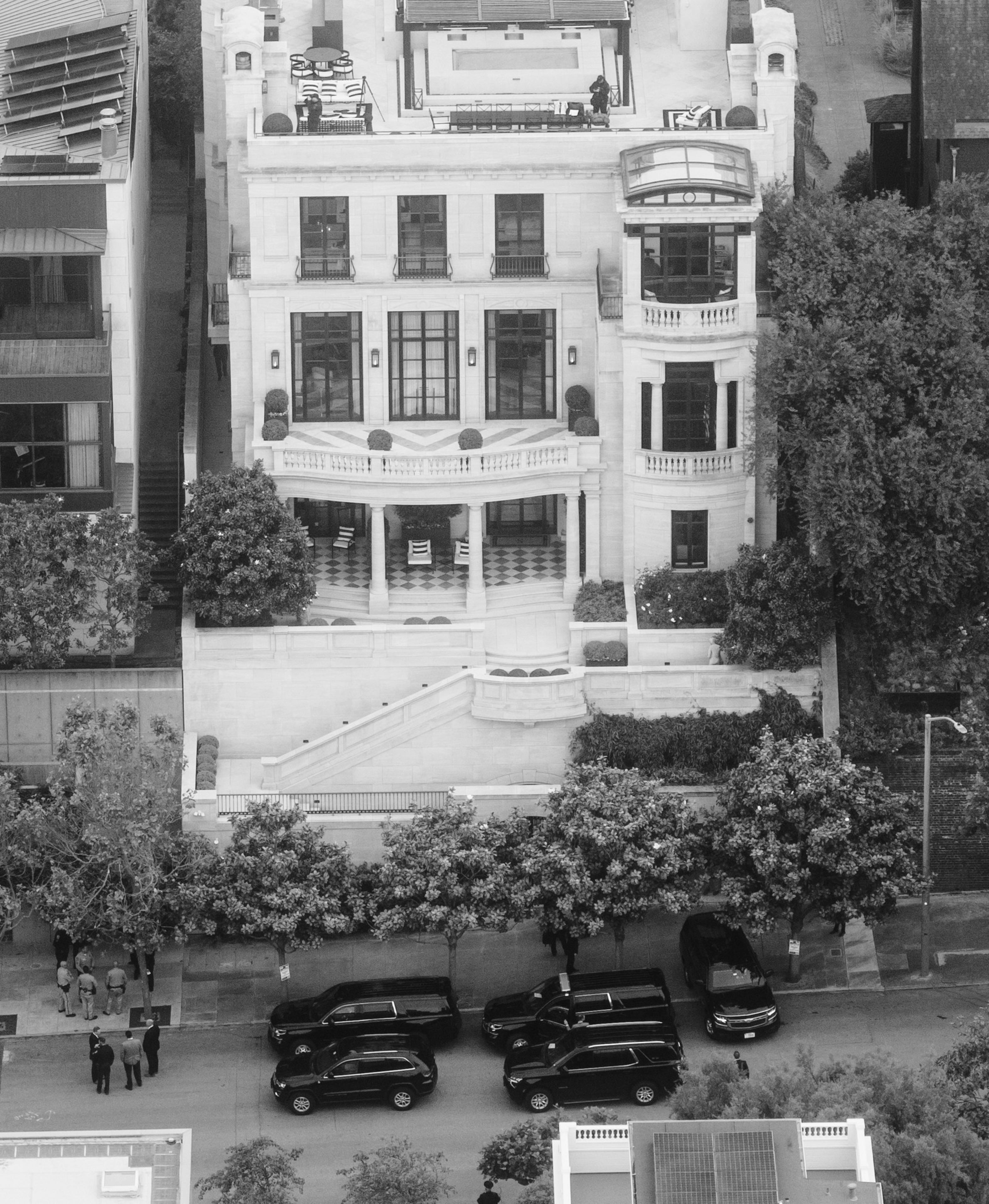 An elegant multi-story building features large windows and a balcony. Black cars park on the street, and a group of people gathers near the trees.