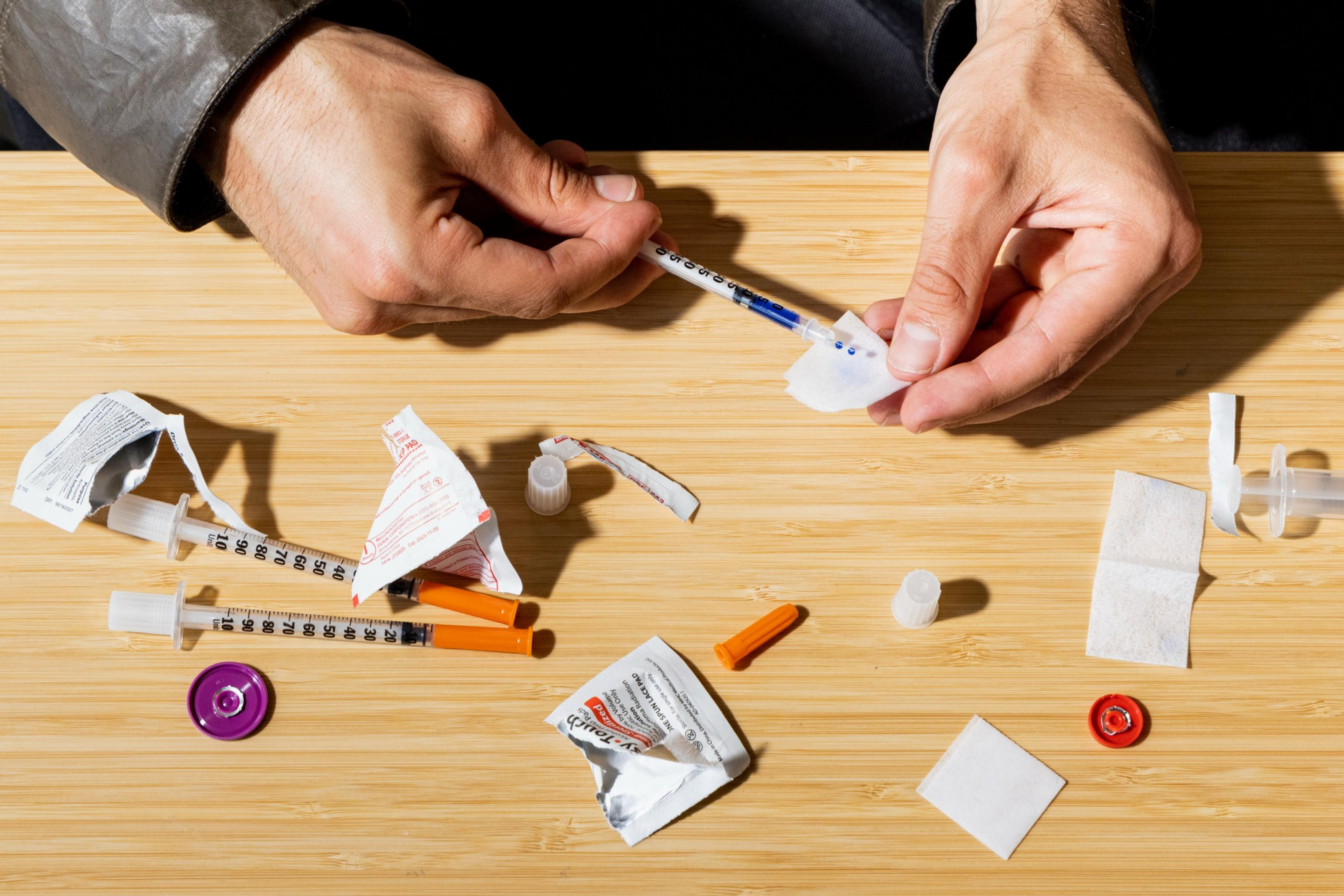Two hands hold a syringe and a small cotton pad above a wooden surface scattered with syringe parts and packaging.