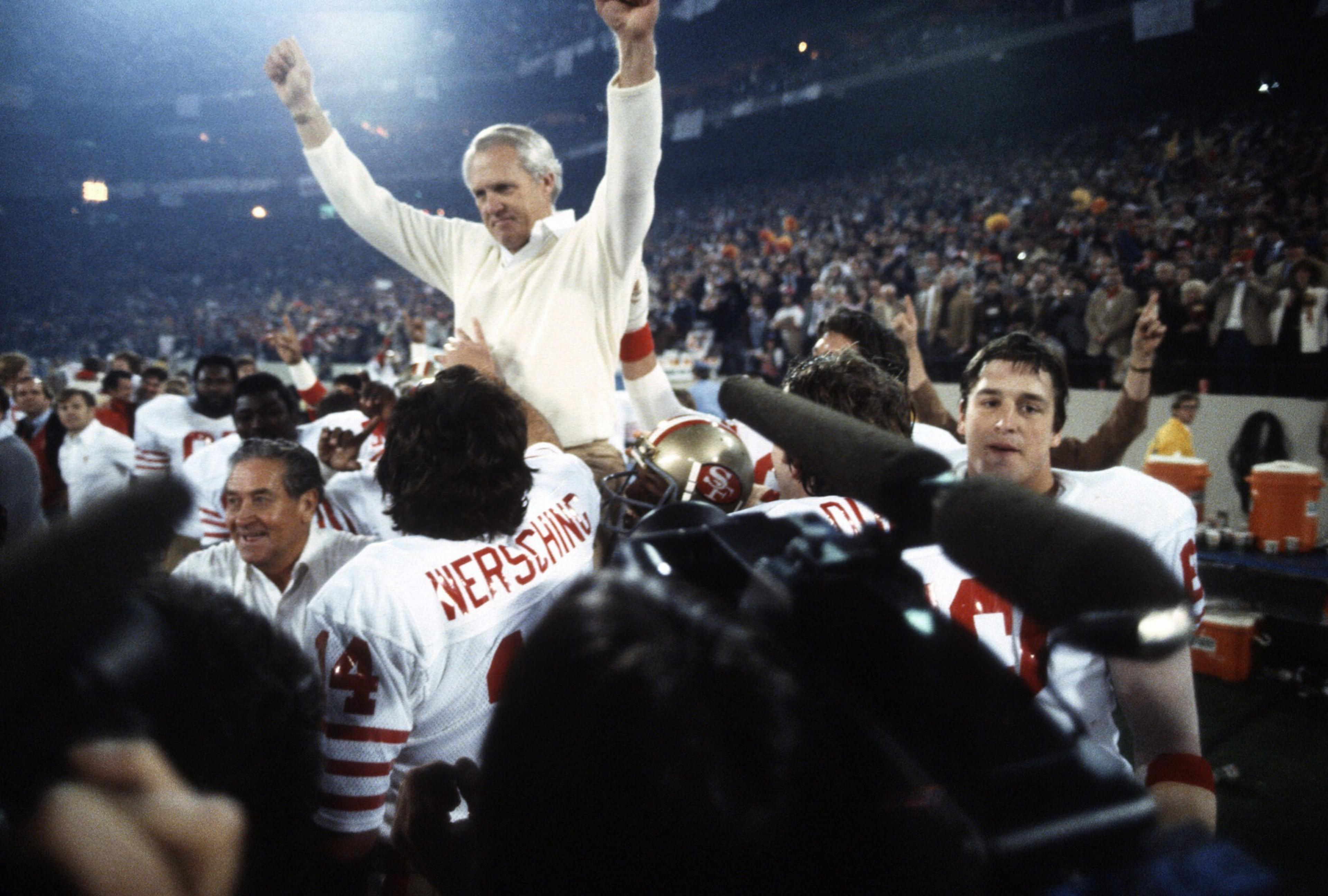 A man in white raises his arms in victory as football players in white and red uniforms celebrate around him in a crowded stadium.