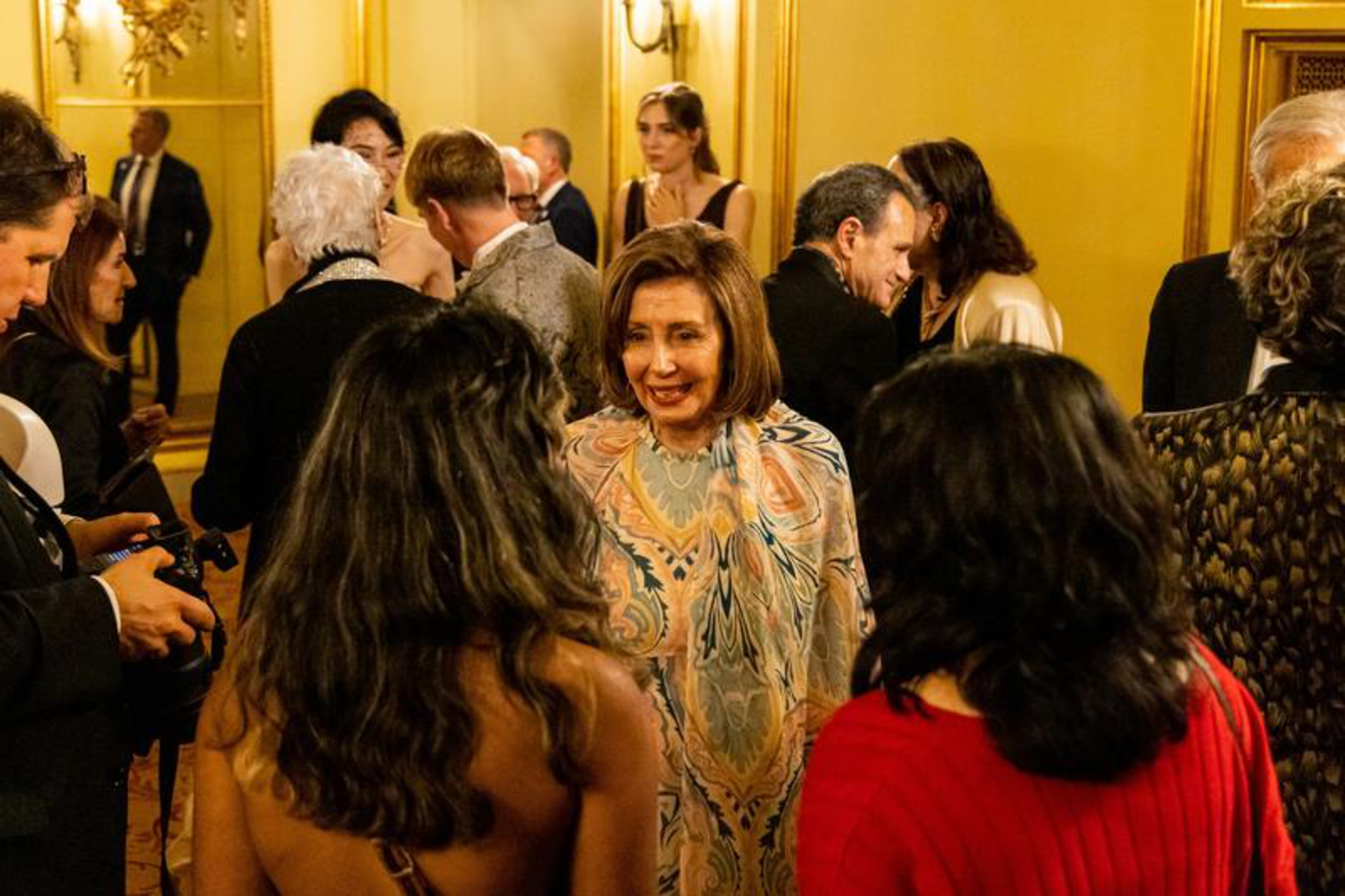 A diverse group of people dressed formally engage in conversation in a warmly lit, ornate room with gold walls and elegant decor.