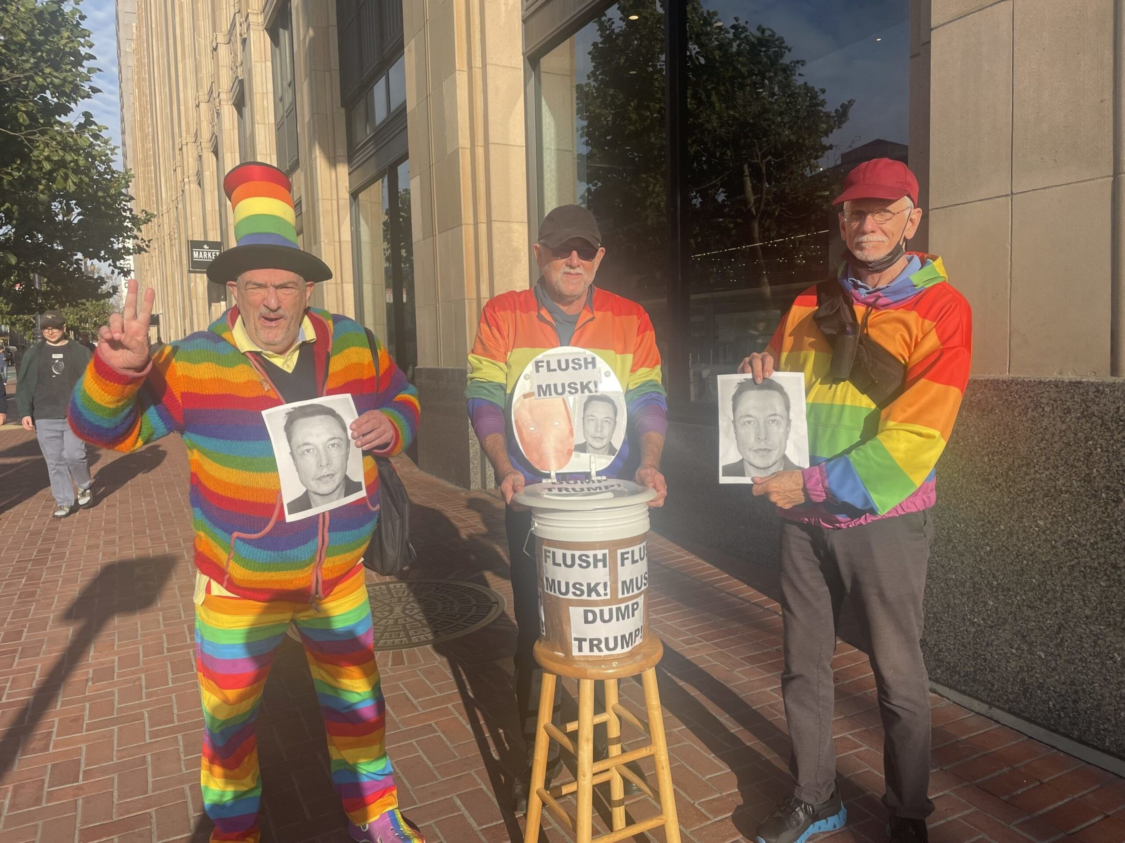 Three men in rainbow clothing hold black-and-white photos of Elon Musk beside a bucket labeled "Flush Musk! Dump Trump" on a wooden stool on a city sidewalk.
