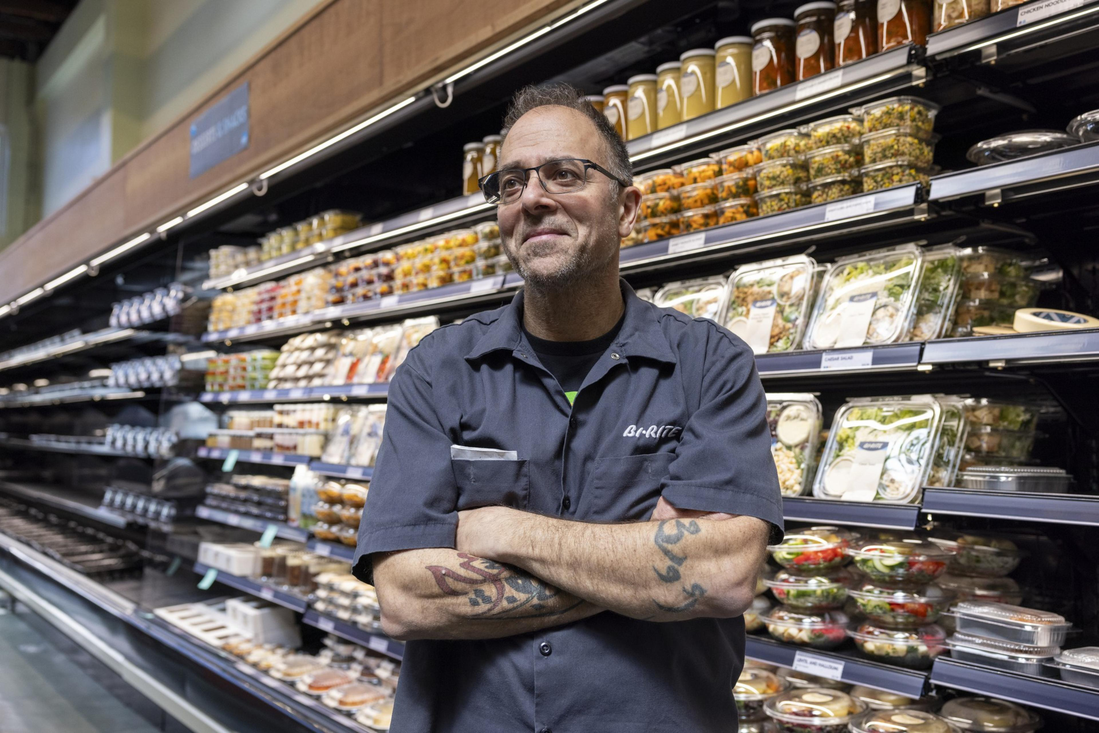 A man with glasses and tattoos stands confidently in a grocery store aisle, with shelves stocked with various packaged foods, including salads and jars in the background.
