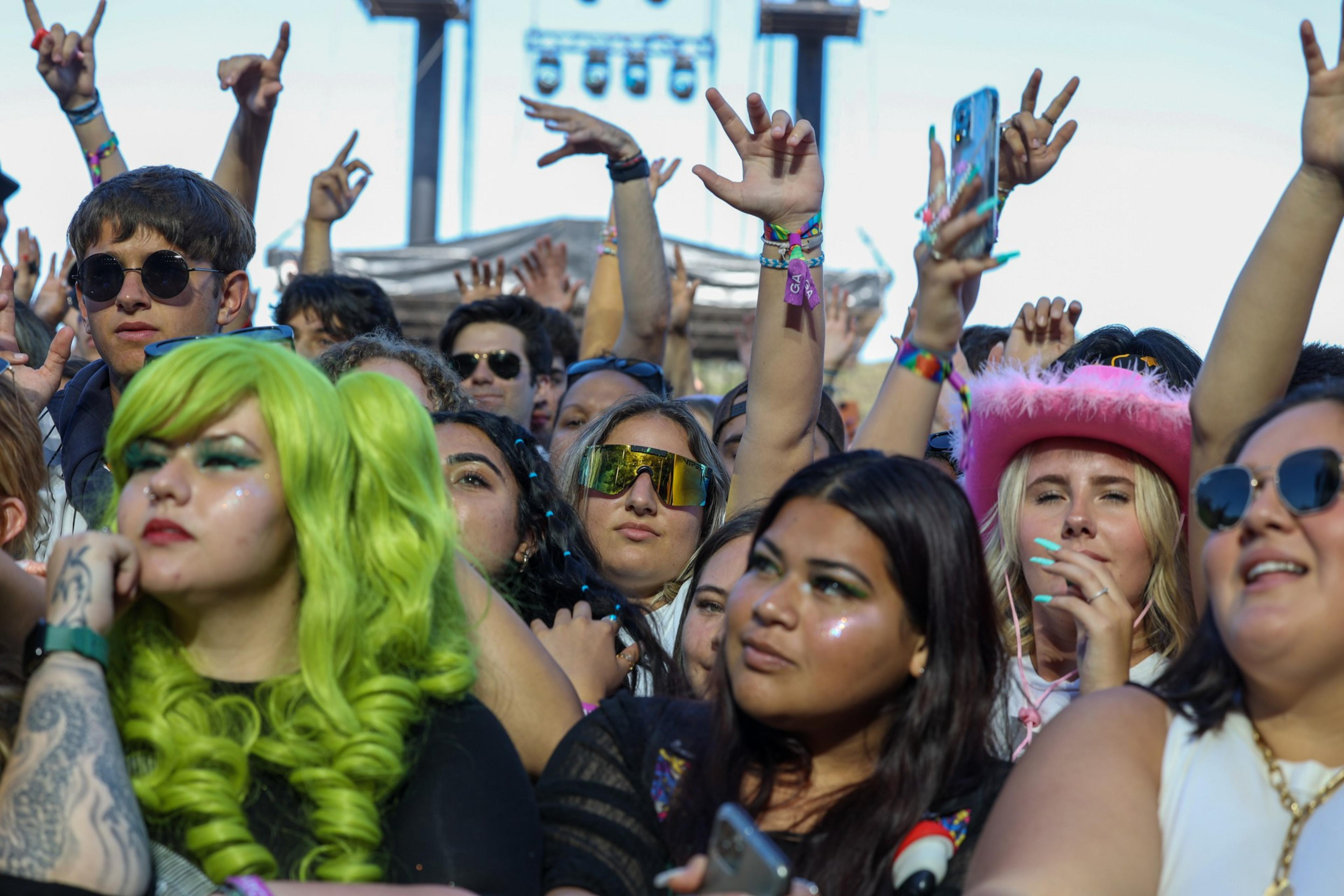 A lively crowd at a festival includes people with colorful hair, sunglasses, wristbands, and a pink cowboy hat, all looking towards a stage.