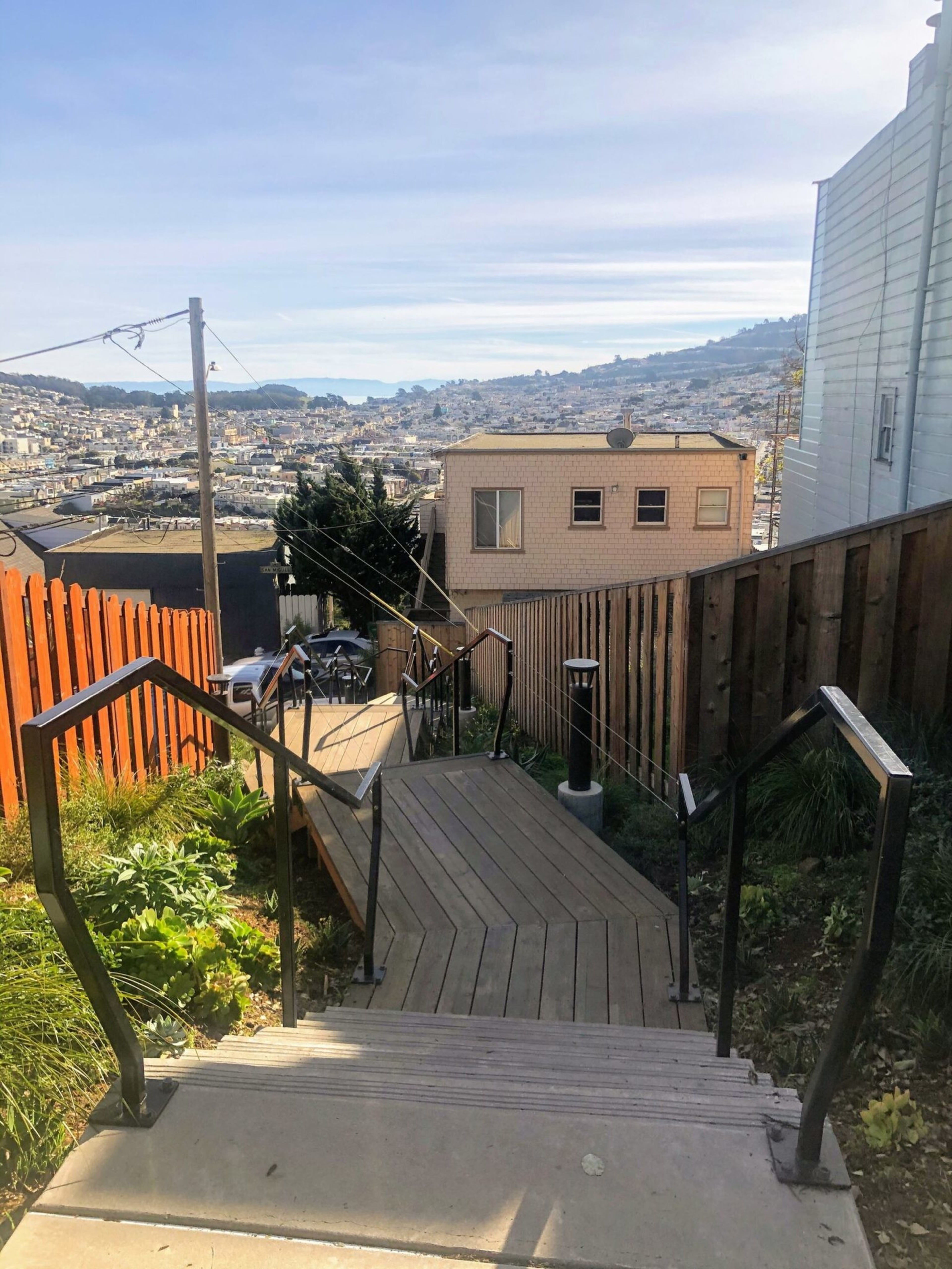 A wooden walkway with metal railings descends between fenced yards, leading toward a cityscape with houses and hills under a clear sky.