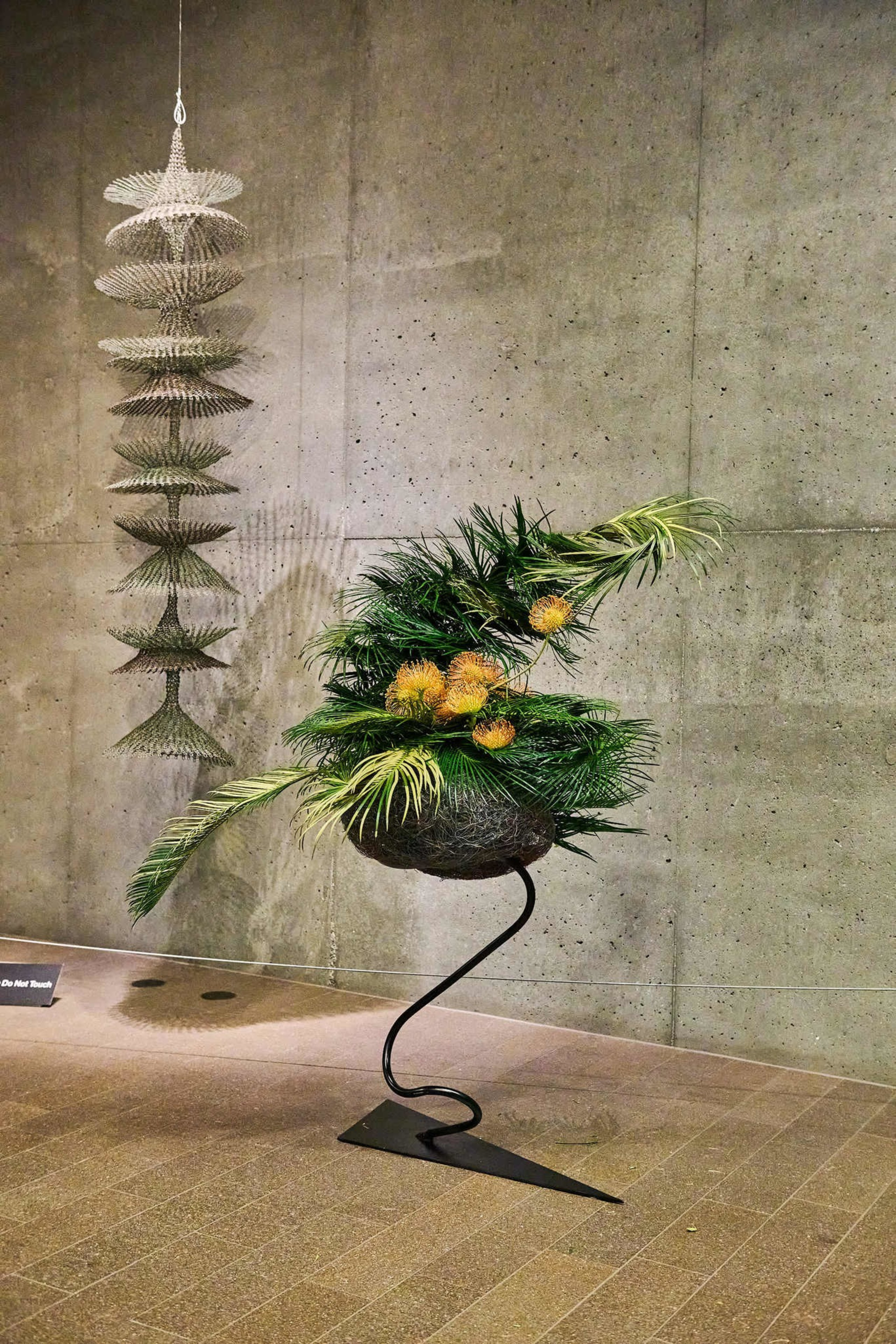The image shows a modern art display in a gallery, featuring a vertical hanging sculpture and an abstract plant arrangement in a wire bowl on a curved stand.