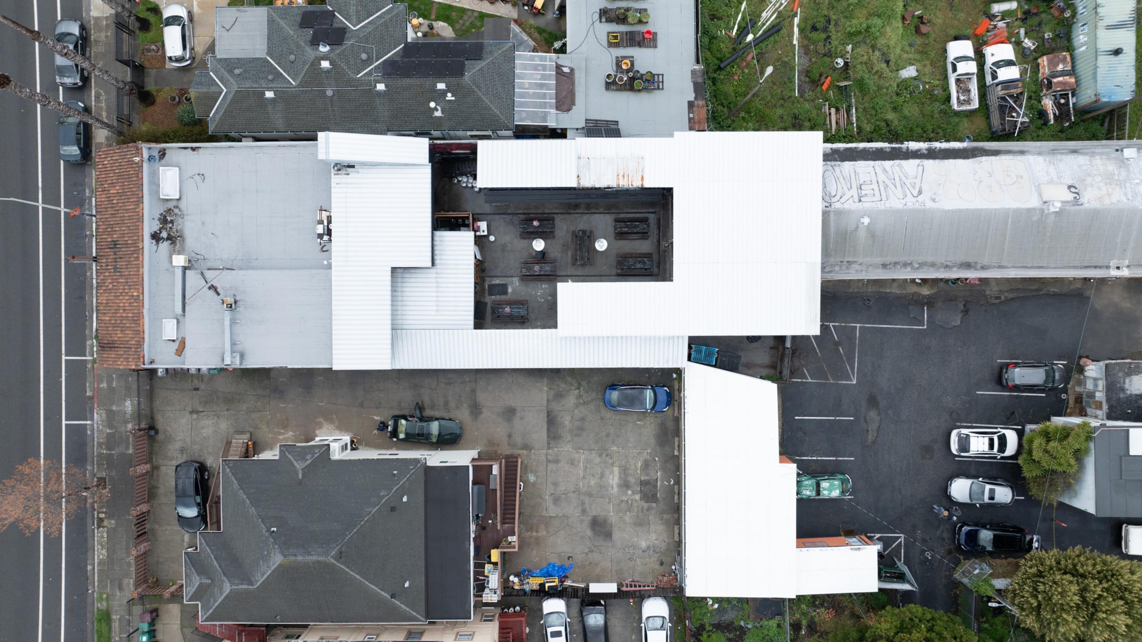 The aerial image shows a cluster of buildings with white and gray roofs, surrounded by parked cars and small patches of greenery.
